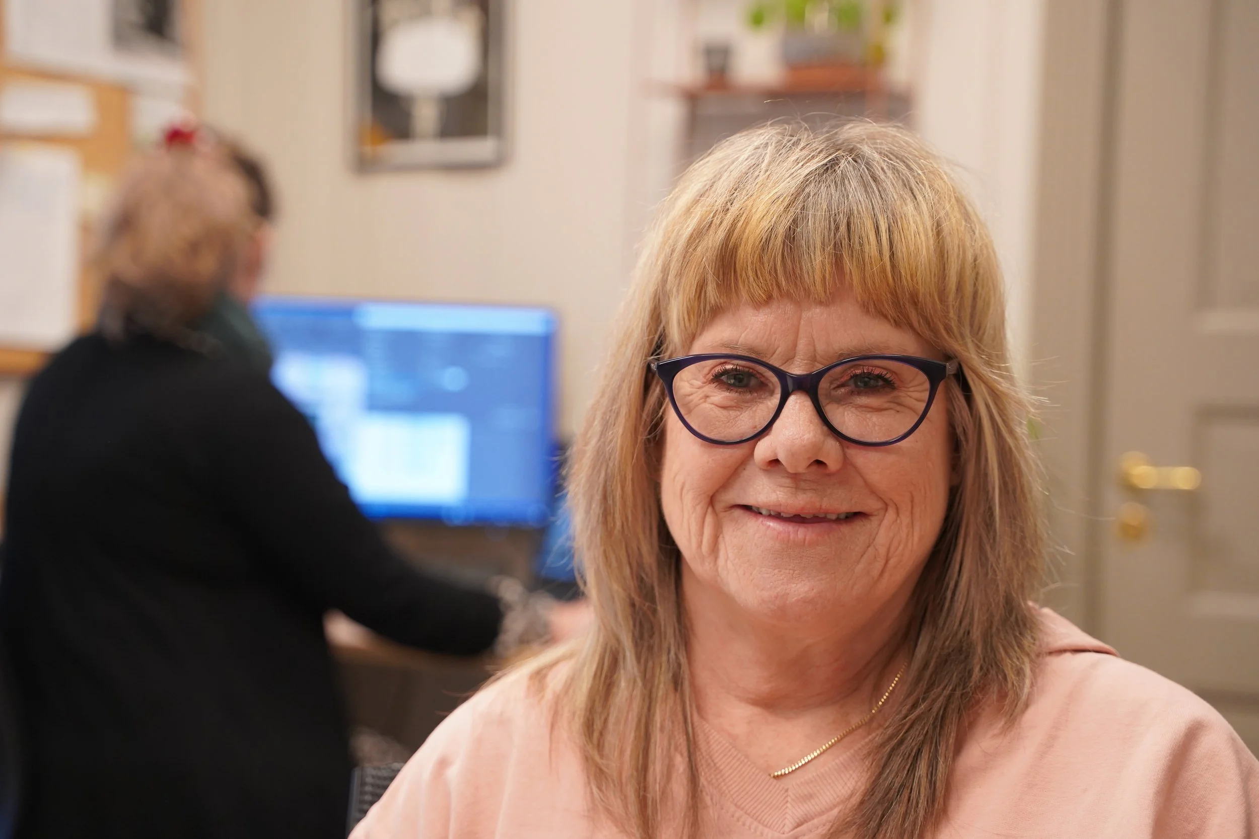 En kvinne med brunt hår og briller smiler mot kamera, mens en annen person jobber foran en dataskjerm i bakgrunnen.