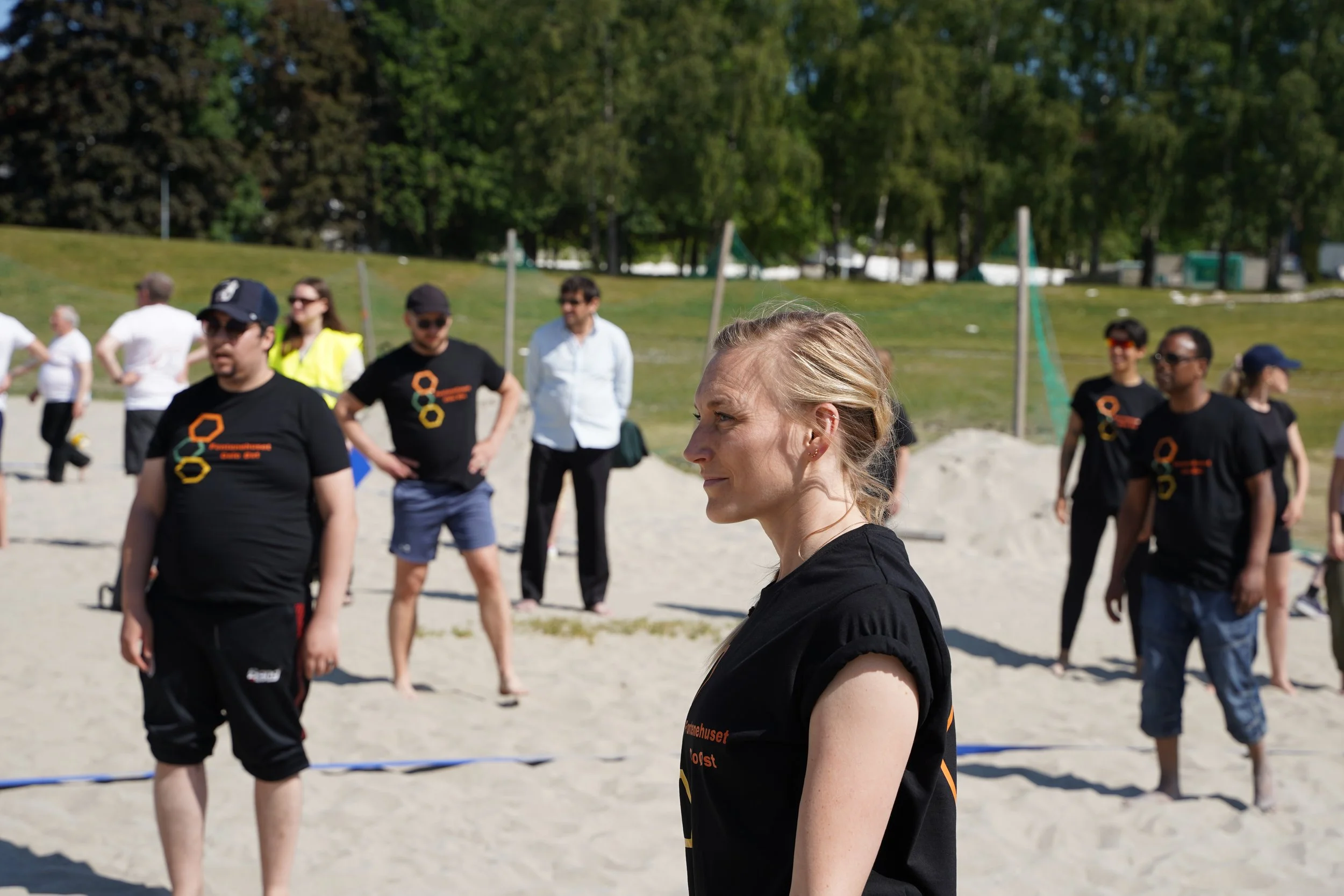 Grupp mennesker på stranden som deltar i en utendørs aktivitet, noen i svarte T-skjorter og noen i casual klær, med trær og grøntområder i bakgrunnen.