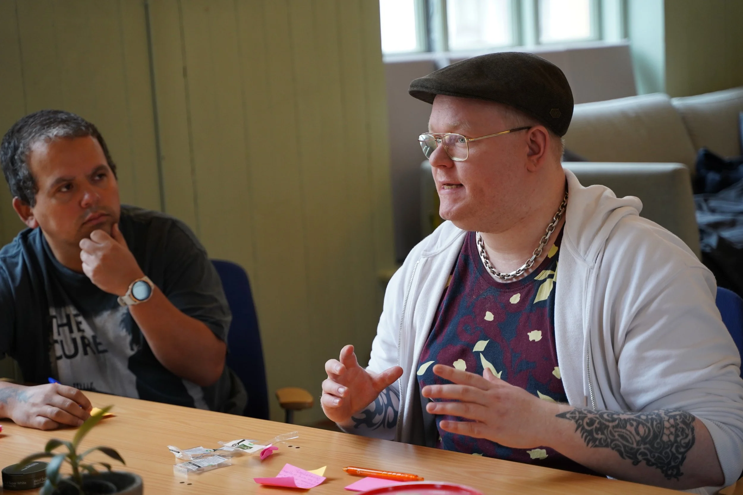 To personer i et møterom som snakker. Den ene personen er kledd i hvit hettegenser, med briller, kjede, mønstrete t-skjorte, caps, og har tatovering på armen. Den andre personen har kort mørkt hår og er i svart t-skjorte, og følger samtalen.
