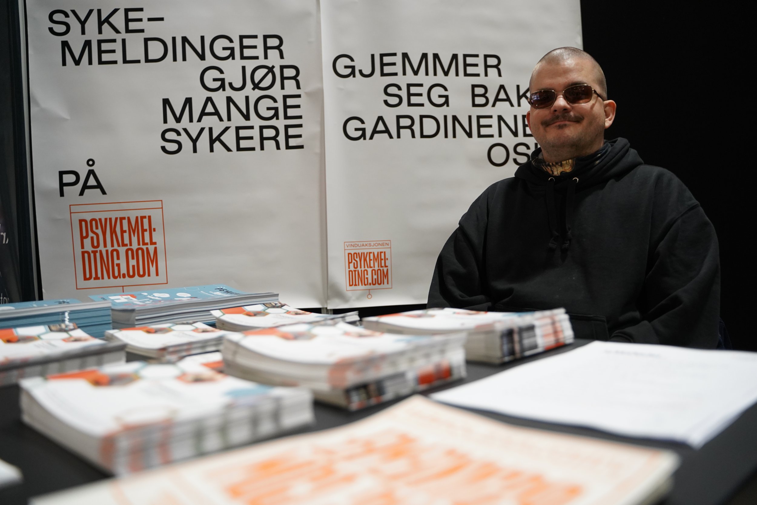 En person sitter ved en stand med brosjyrer og plakater, bak en stor plakat med teksten "SYKEMELDINGER GJØR MANGE SYKERE" og annonse for psykemeldingsnettstedet Psykemeding.com.