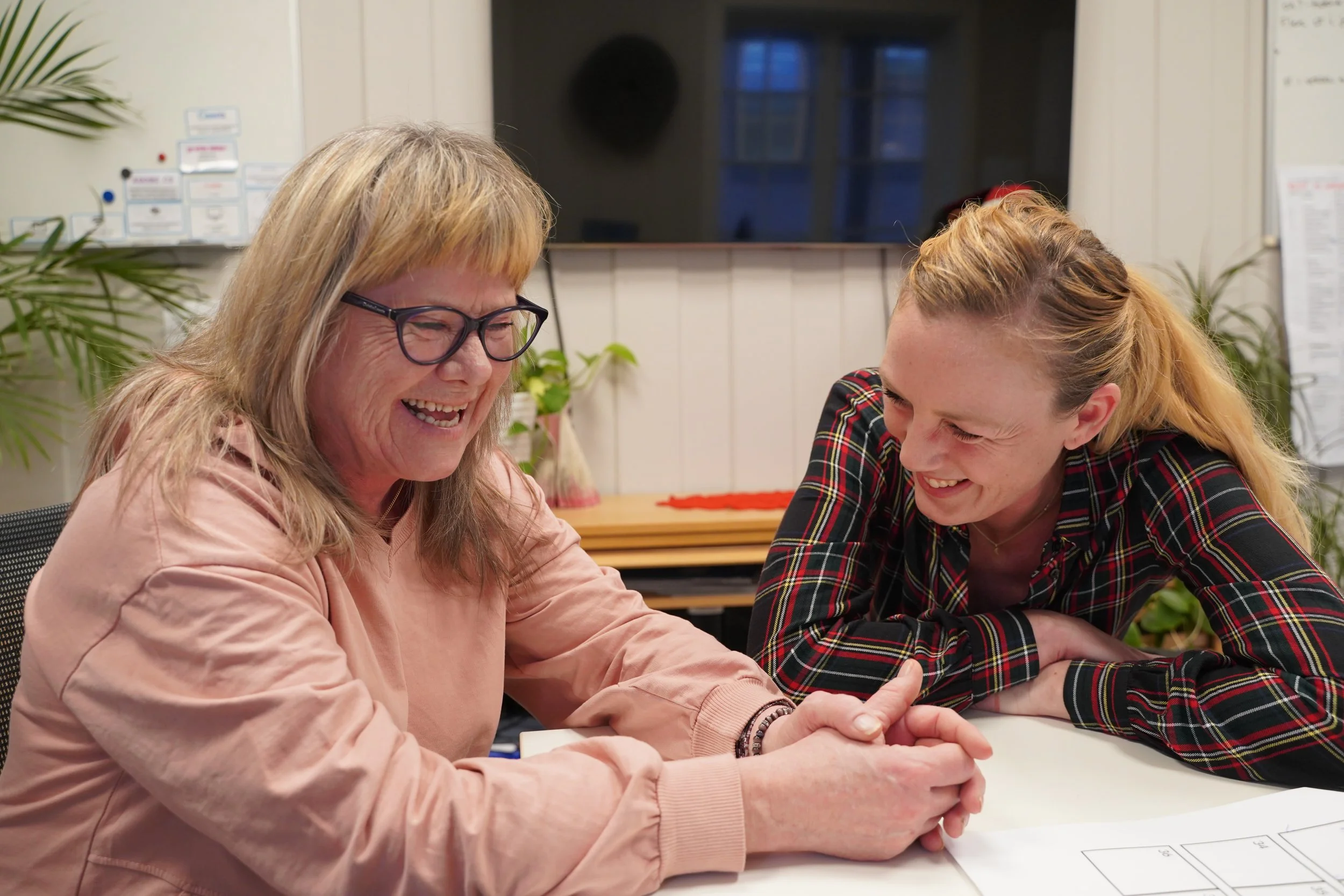 To kvinner smiler og ler mens de har en prat ved et bord, en av dem har på seg briller og rosa genser, den andre har på seg rutete skjorte.
