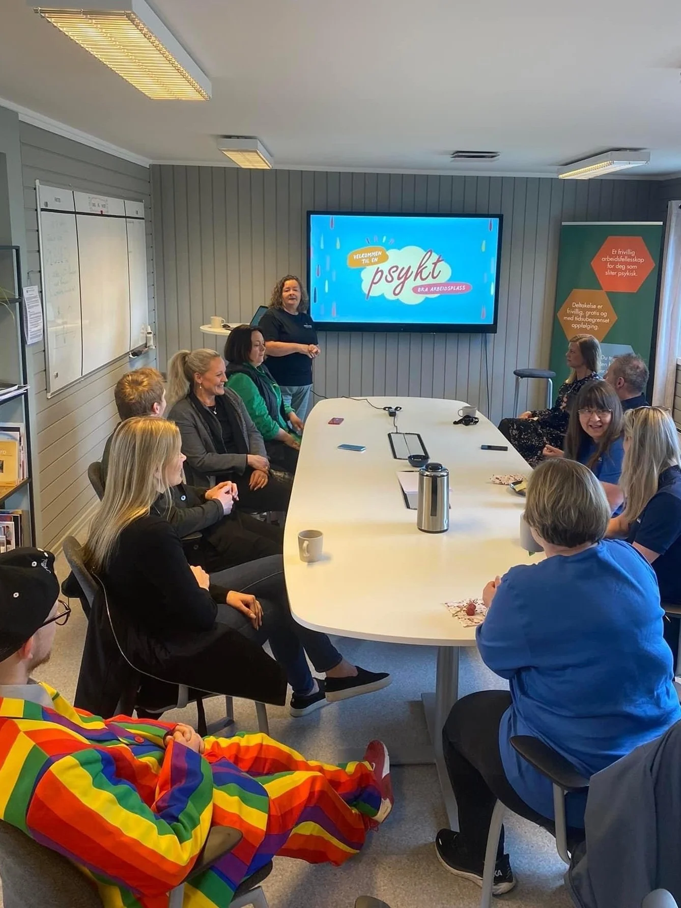 En grupp mennesker deltar i et foredrag eller en workshop i et møterom, med en skjerm som viser teksten "Velkommen til en psykt" og flere personer sitter rundt et langbord.