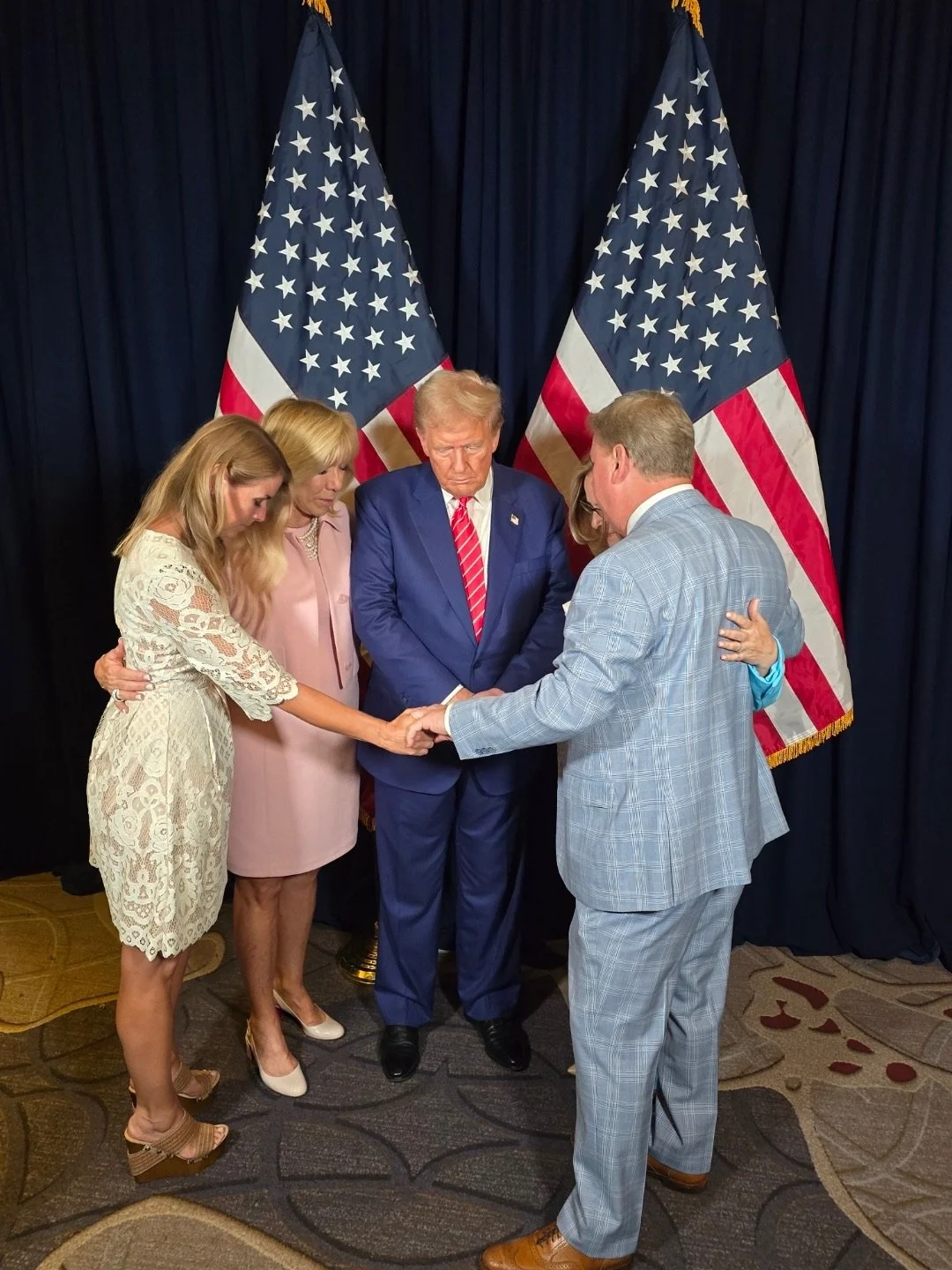 A group of five people, including Donald Trump, holding hands and praying with heads bowed, in front of two American flags and a dark curtain.