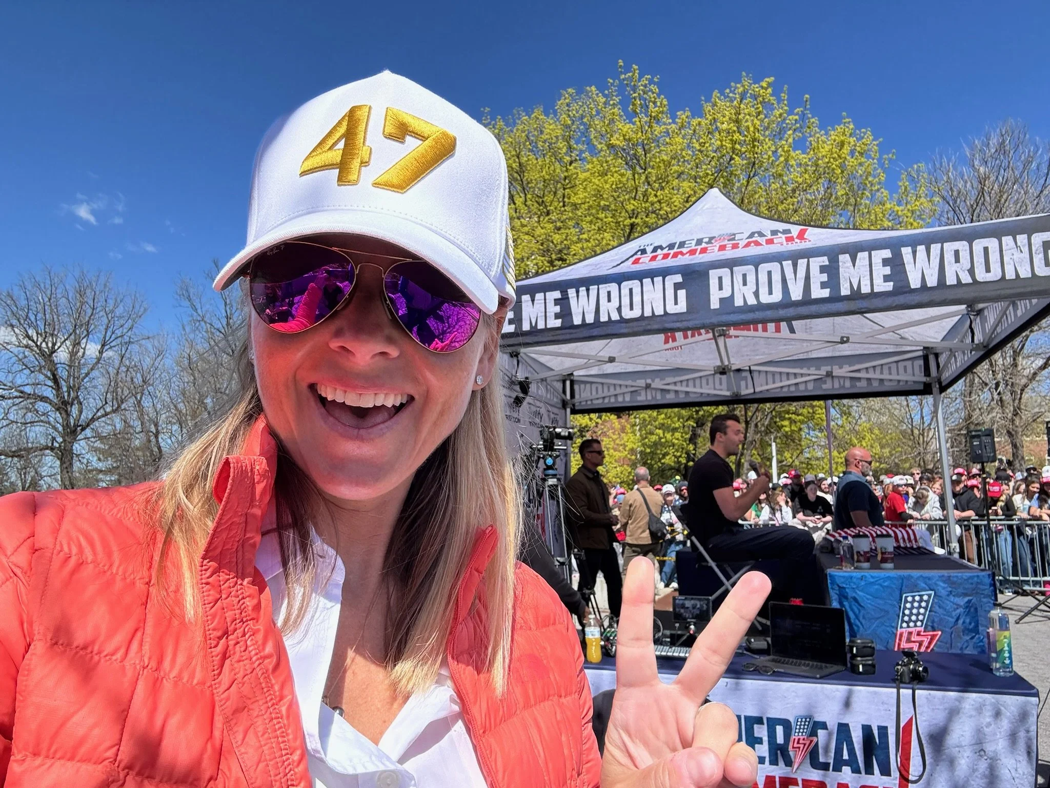 A smiling woman wearing a white cap with the number 47, purple sunglasses, and an orange jacket taking a selfie at an outdoor event with a crowd and a stage in the background. The stage has a booth with a banner reading 'PROVE ME WRONG' and 'AMERICAN