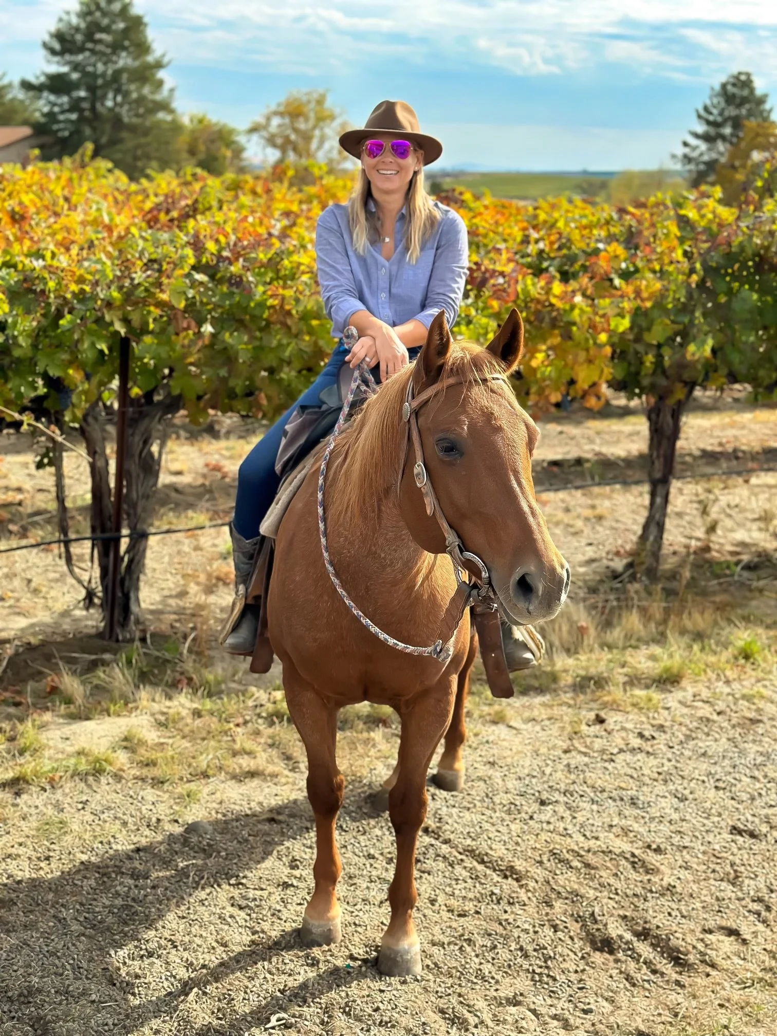A woman riding a horse in a vineyard during autumn. She is wearing a wide-brimmed hat, pink sunglasses, a blue button-up shirt, and jeans, smiling at the camera.
