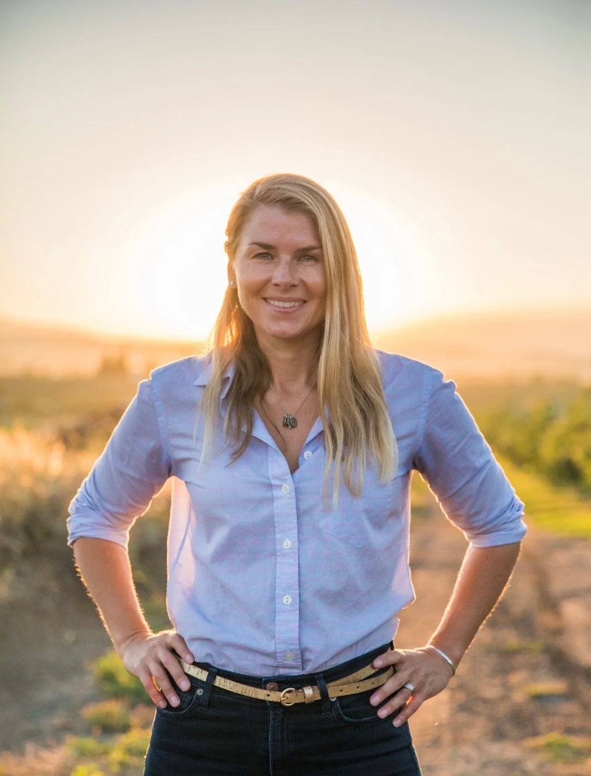 A woman with blonde hair wearing a light blue button-up shirt and black pants standing outdoors at sunset, smiling with her hands on her hips.