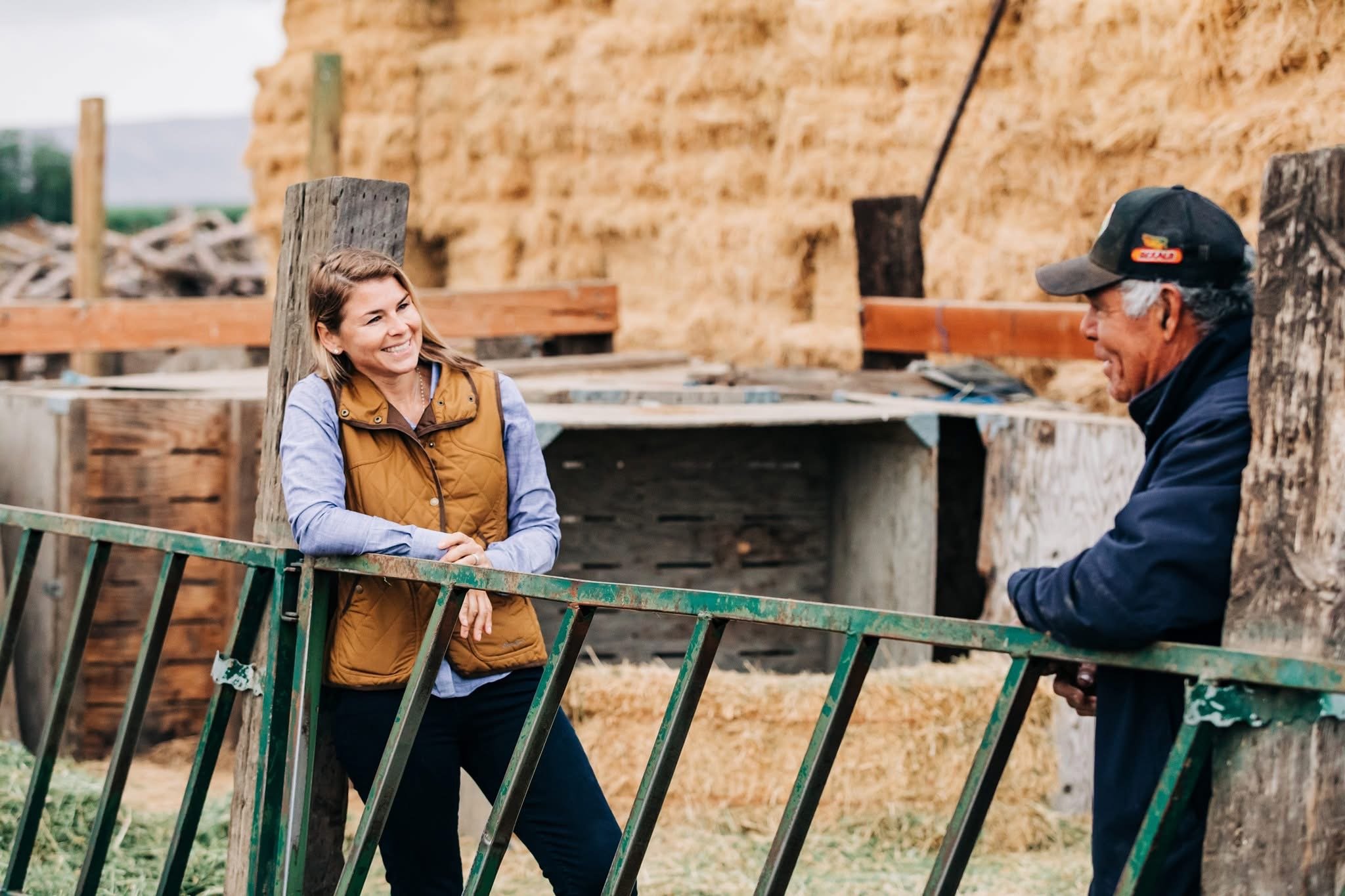 A woman and a man smiling and talking outdoors near a wooden fence and rustic building.