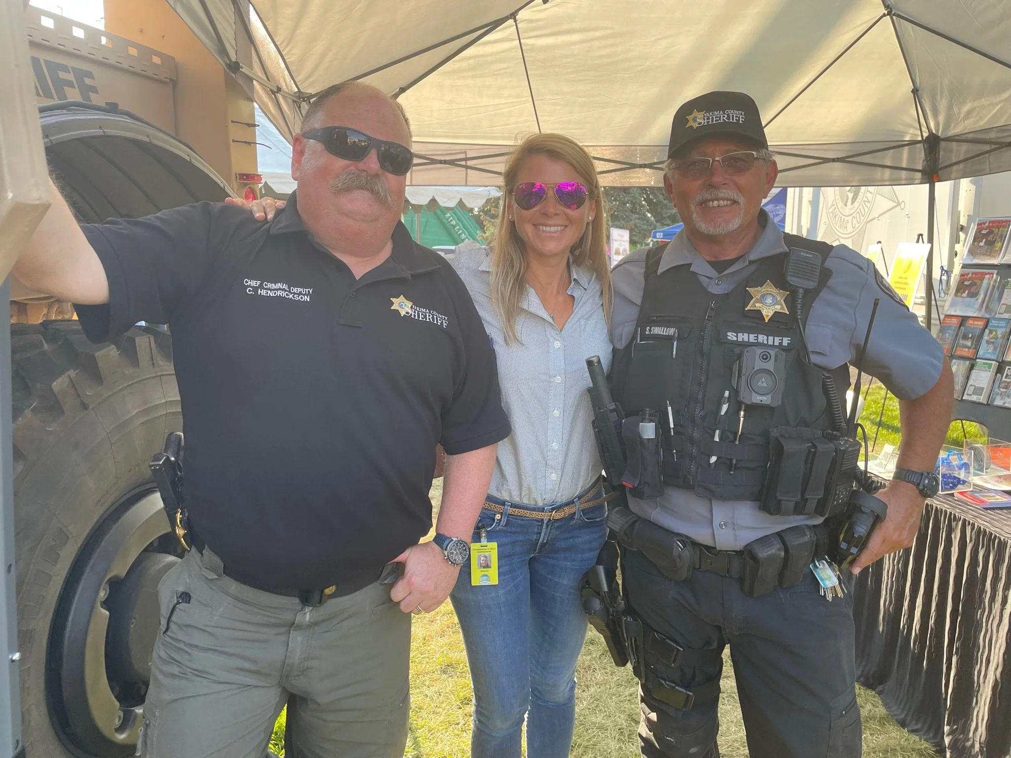 Three smiling Caucasian people, two men and one woman, standing under a canopy at an outdoor event. The man on the left is wearing a black shirt with 