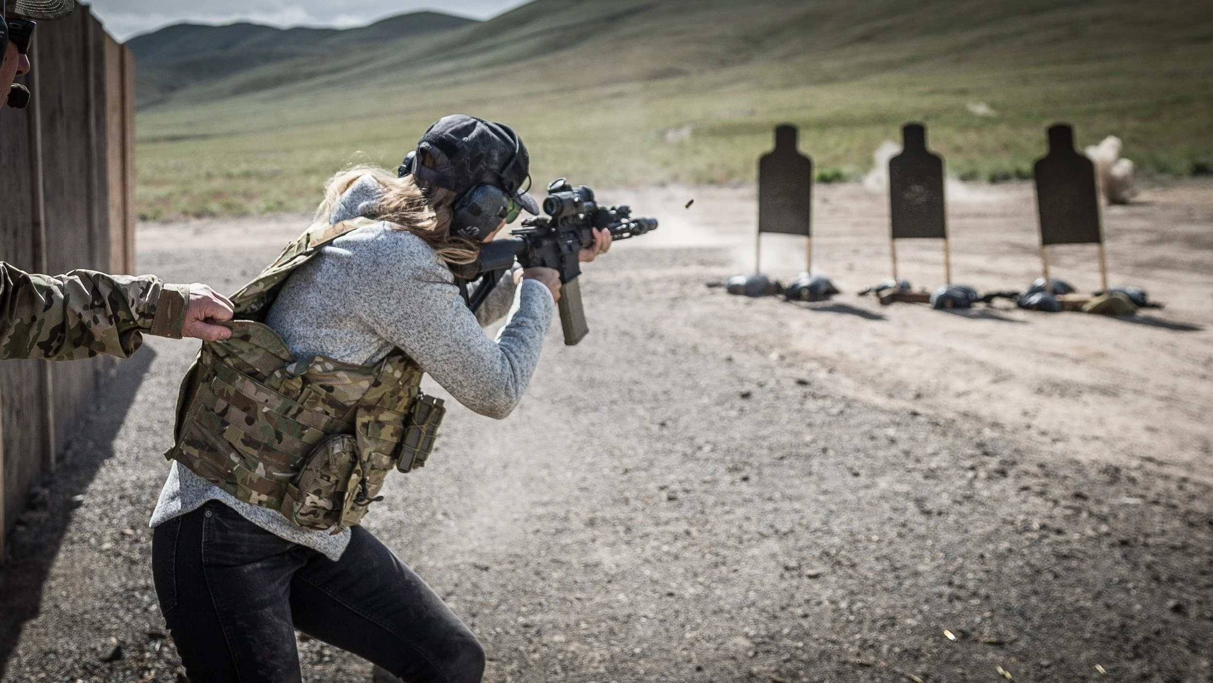 A woman in camouflage tactical gear aiming a rifle at target silhouettes during firearm training at outdoor shooting range.