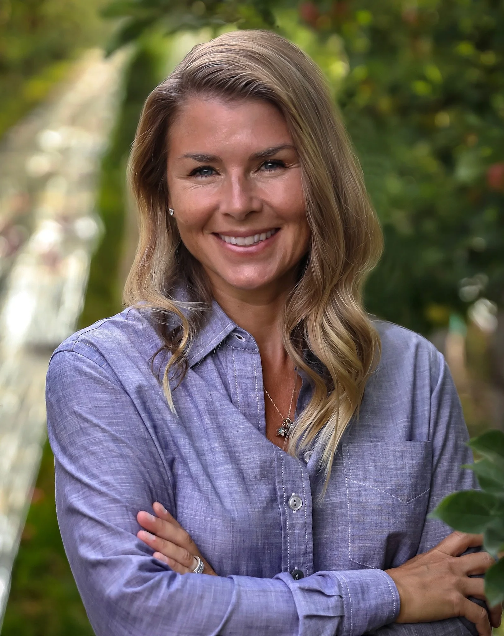 A smiling woman with blonde hair wearing a button-up shirt stands outdoors with her arms crossed, surrounded by blurred green trees.