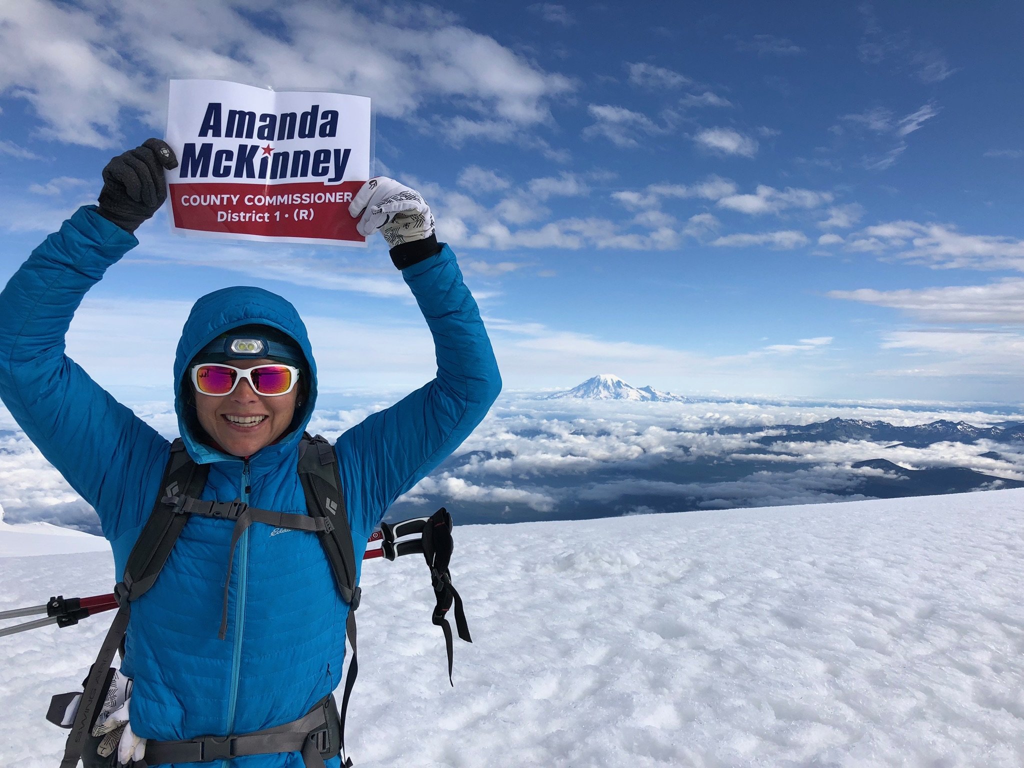 A woman dressed in blue winter gear with sunglasses and a headlamp, smiling and holding a campaign sign that reads 'Amanda McKinney, County Commissioner, District 1 (R)', on snowy mountain summit with a volcano in the background, under a partly cloud