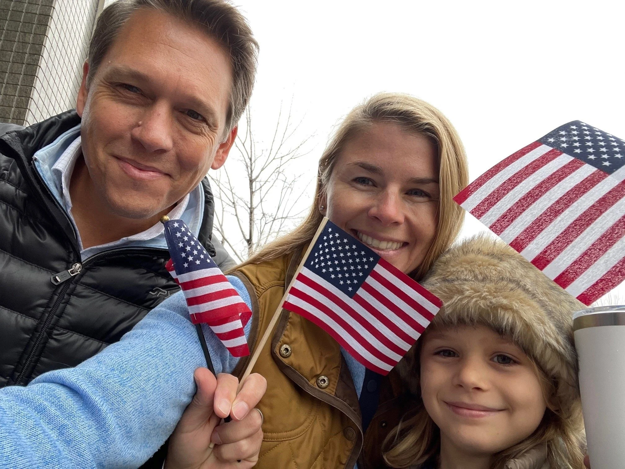 A family of three smiling outdoors, holding small American flags. The father is on the left, the mother in the middle, and the child on the right wearing a fur-lined hood. Overcast sky, bare trees in the background.