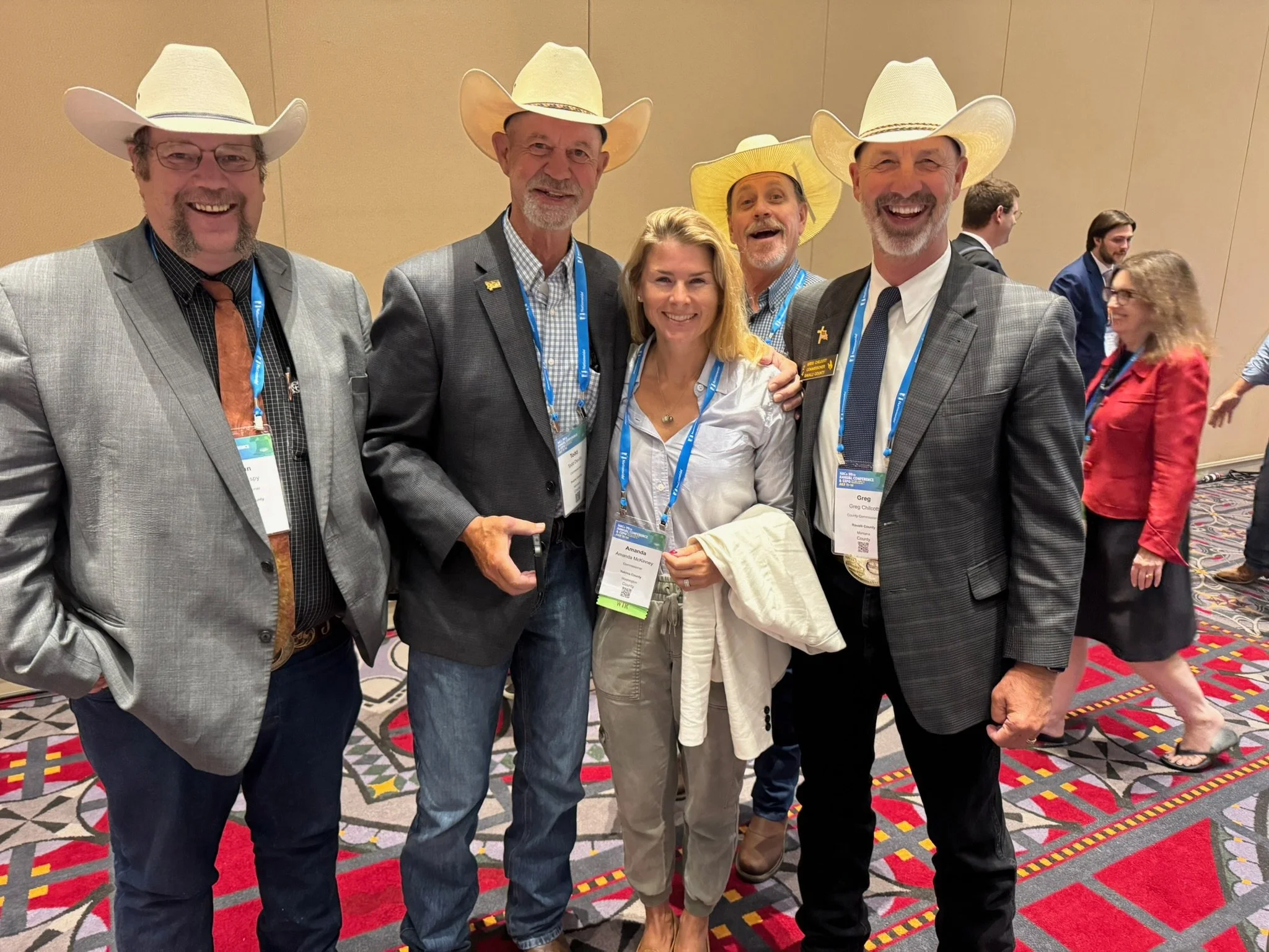 Group of five people, four men and one woman, wearing cowboy hats and business attire, standing together and smiling at an indoor event.
