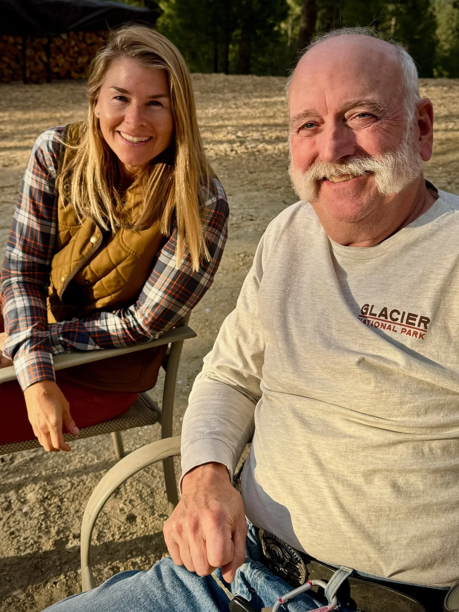 A young woman and an older man with a white beard sitting outside in a natural setting, smiling at the camera.