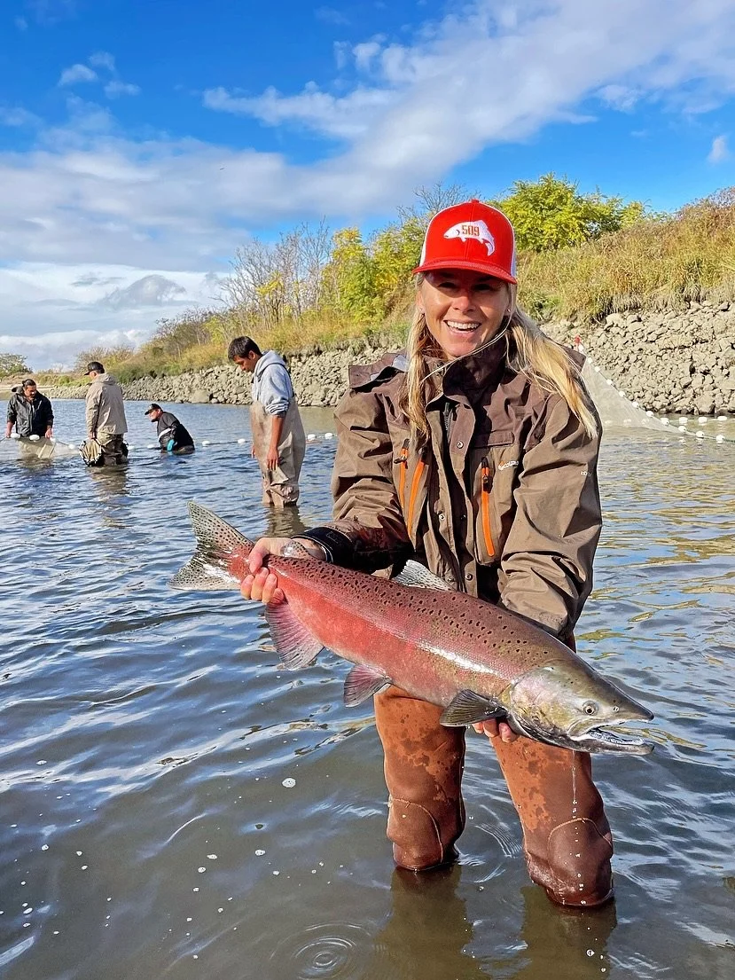A woman in a brown jacket and red hat smiling while holding a large rainbow trout in a river, with other people fishing in the background and trees on the riverbank under a cloudy sky.