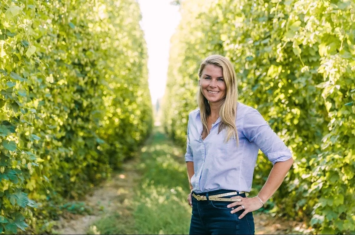 A smiling woman standing in a lush green vineyard with rows of grapevines on either side.