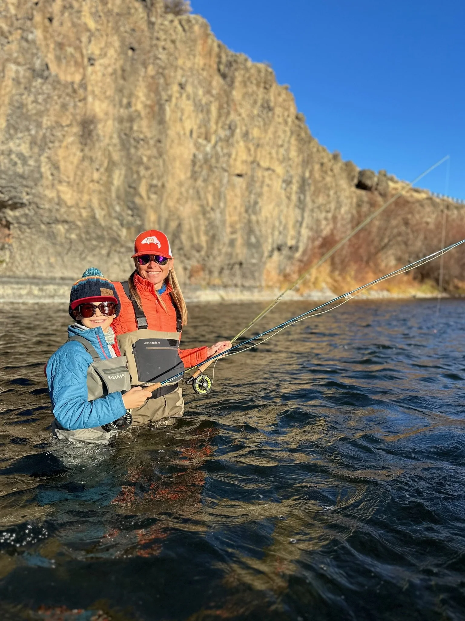 A woman and a child fishing in the water, both wearing life jackets, hats, and sunglasses with a rocky cliff and blue sky in the background.