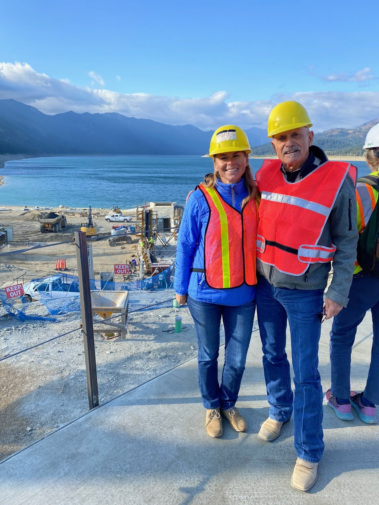 Two people in hard hats and safety vests standing on a concrete path near a construction site with a lake and mountains in the background.