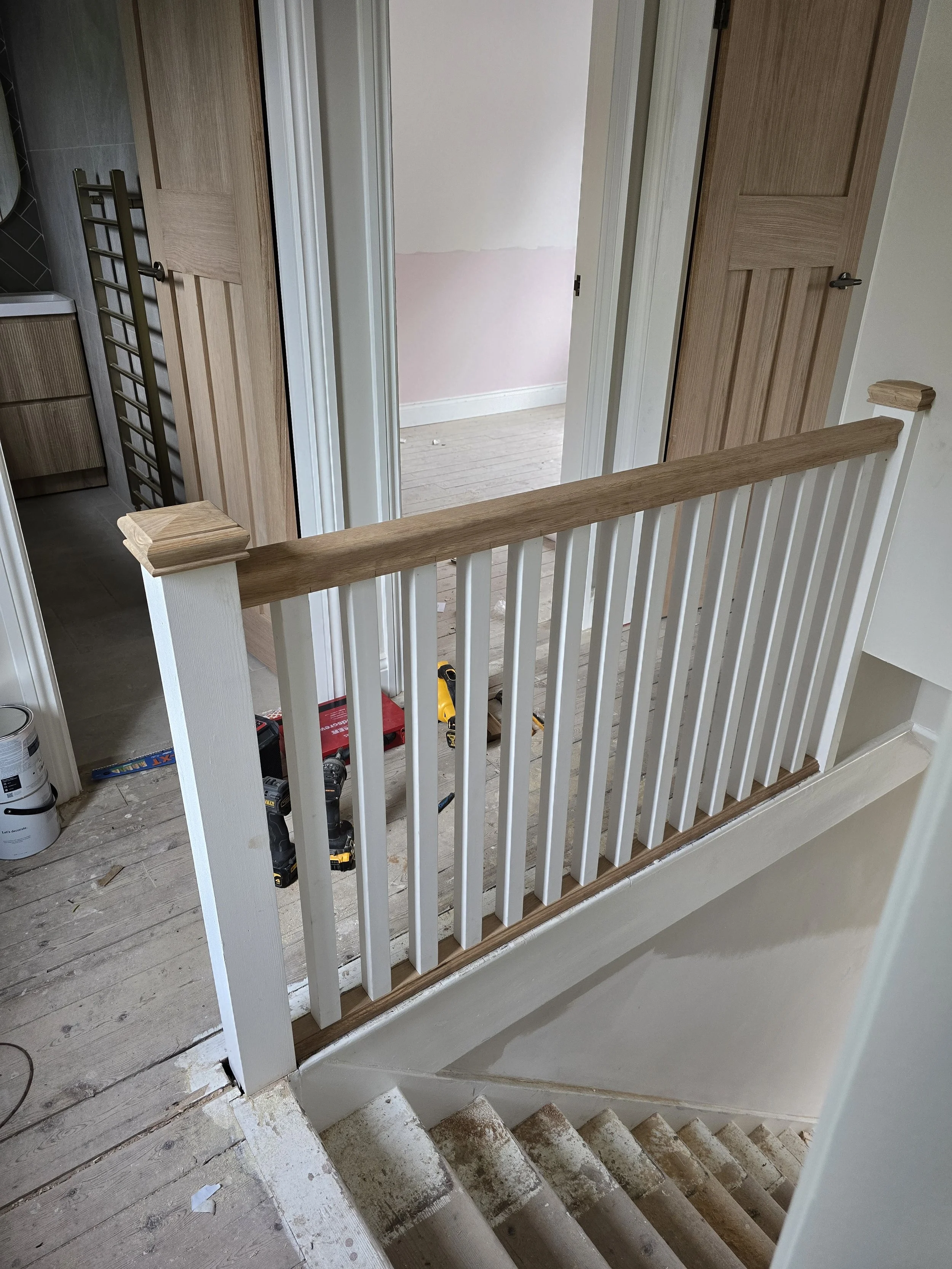 Construction site on a staircase landing, with tools and paint cans visible, railing partially painted, and bedrooms with closed doors.