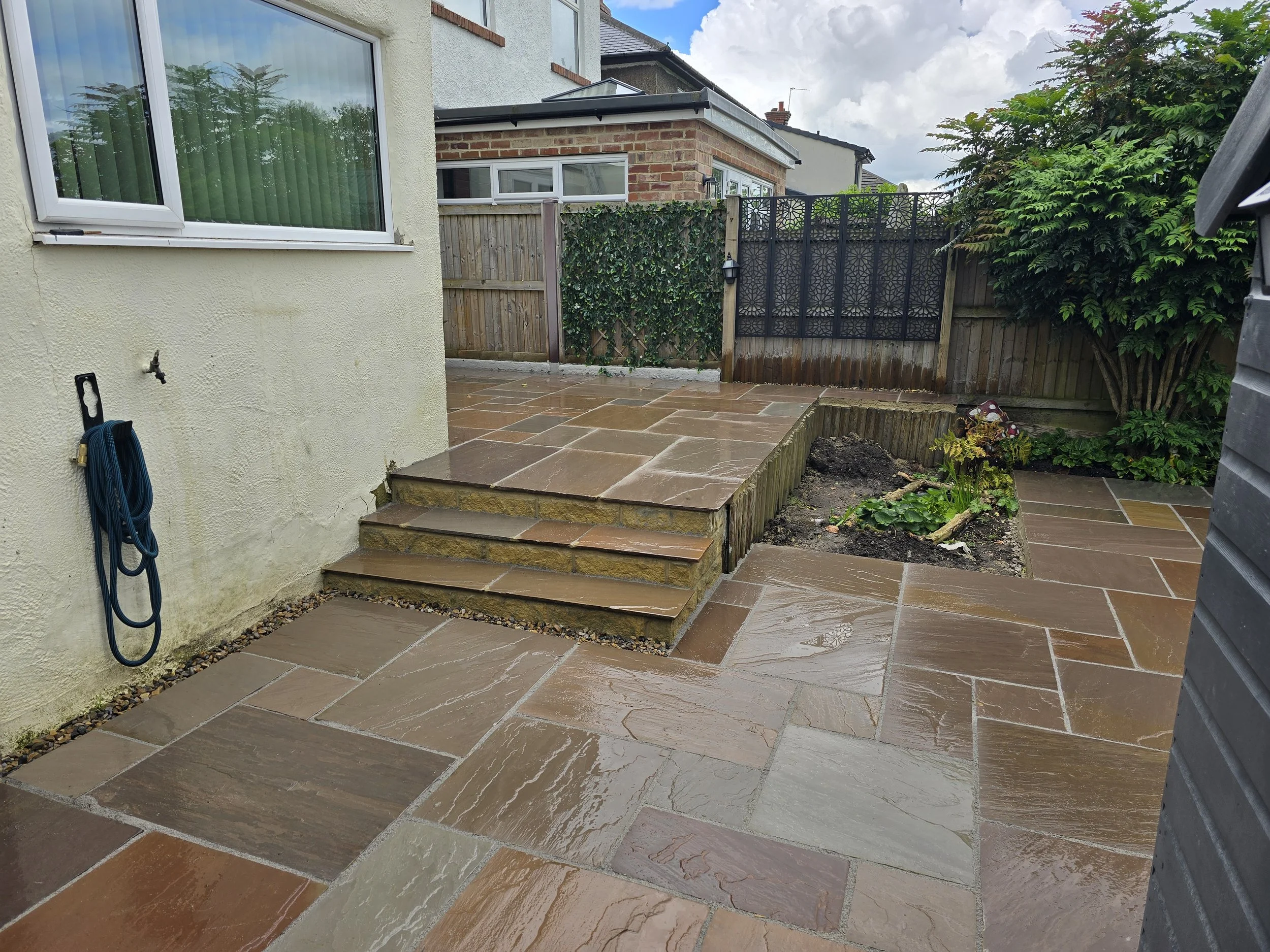 Clean, wet stone patio with a small raised garden bed and steps leading to an elevated section, backyard with wooden fences, and house walls with a large window and garden hose.