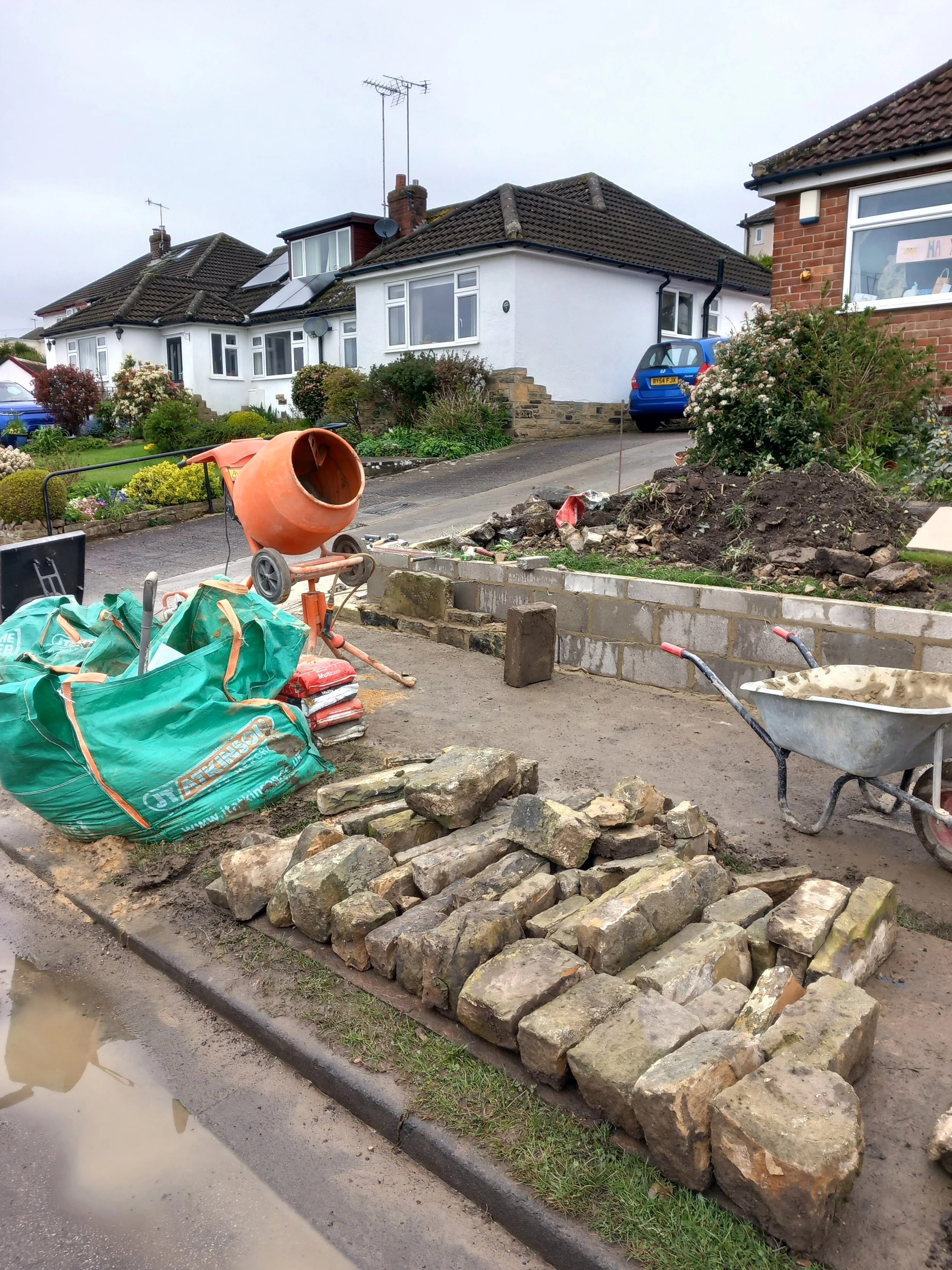 Front yard under construction with a pile of bricks, a cement mixer, bags of cement, a wheelbarrow, and various construction tools. Residential houses with gardens and parked cars are visible in the background.