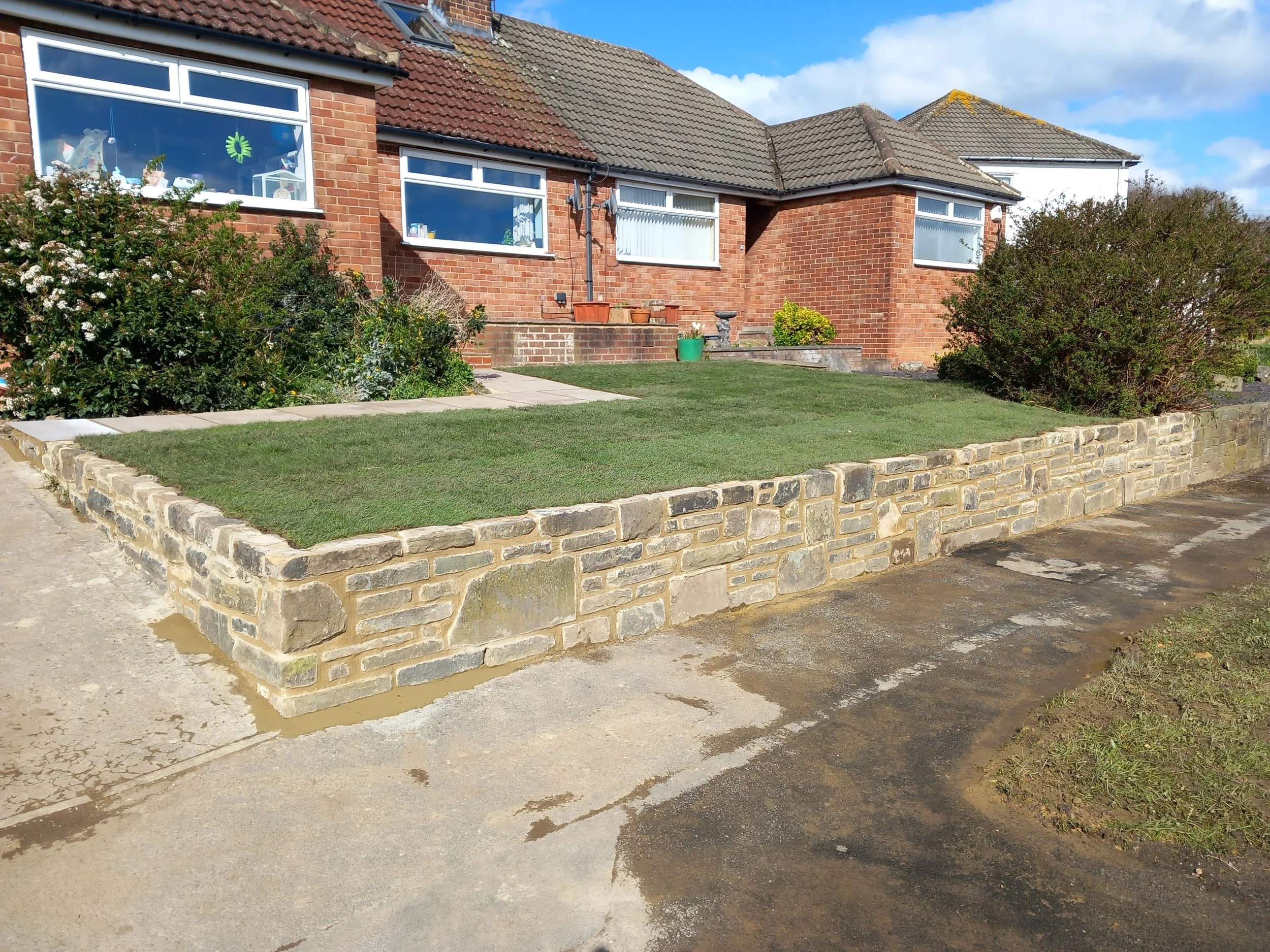 Front yard with a brick house, green lawn, landscaped bushes, flower pots, and a stone wall edging the garden.