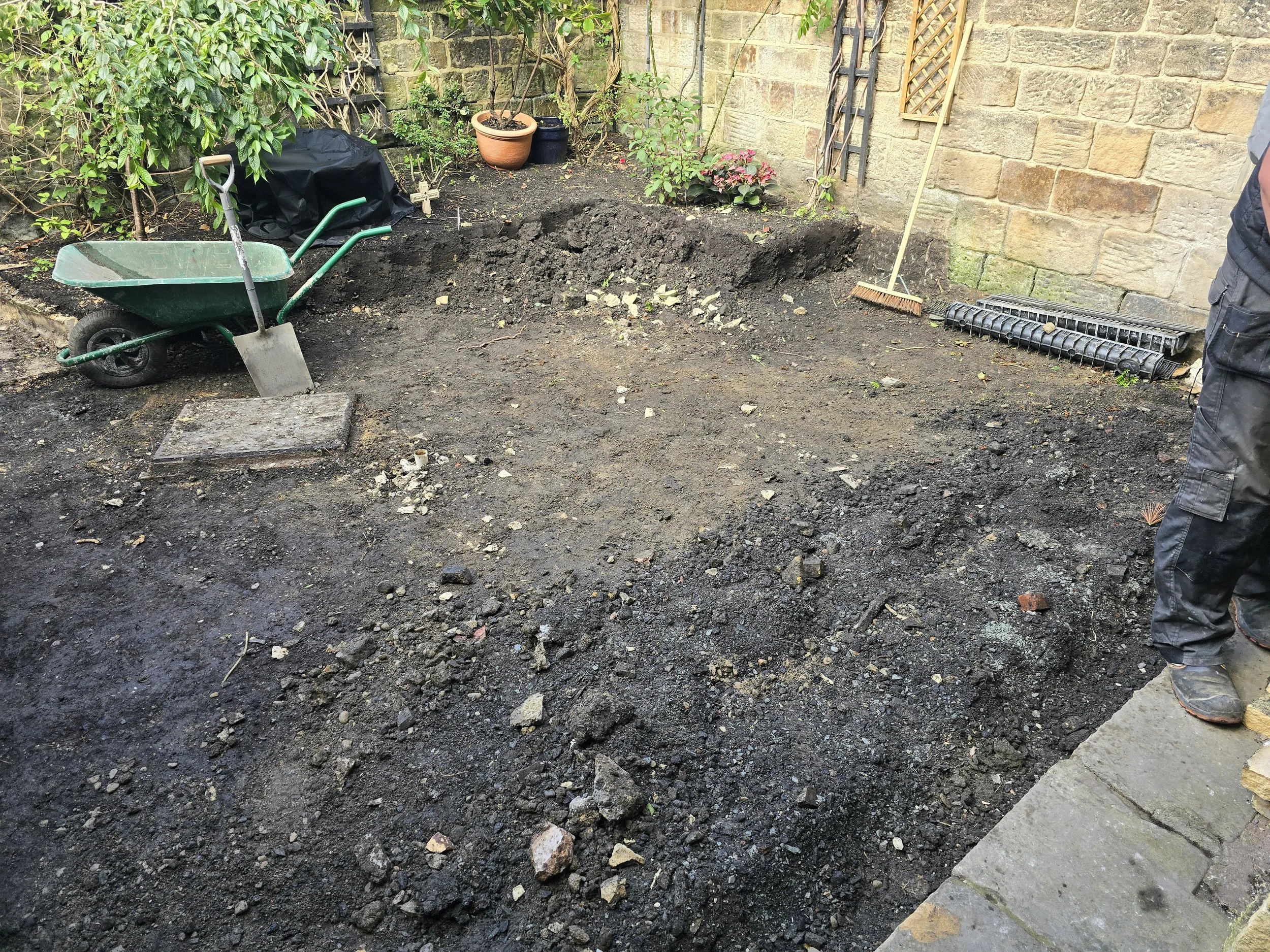 A backyard garden area being prepared for planting, with dark soil spread out, a wheelbarrow, a small shovel, a rake, potted plants, and garden edging tools visible.
