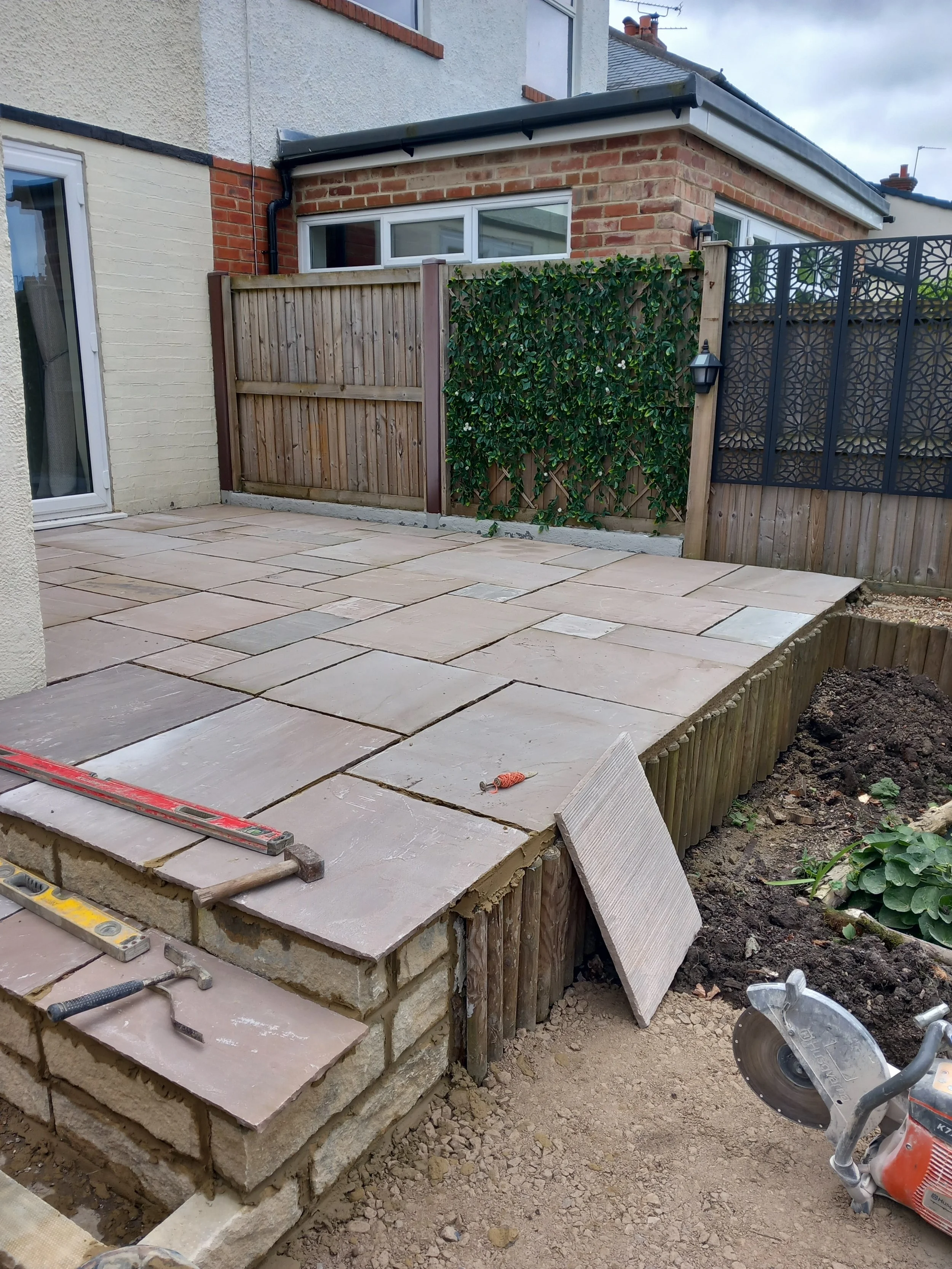 A backyard patio under construction with paving stones, a partially built stone wall, and various tools, including a saw and a level, with a wooden fence, green plants, and a brick and stucco house in the background.