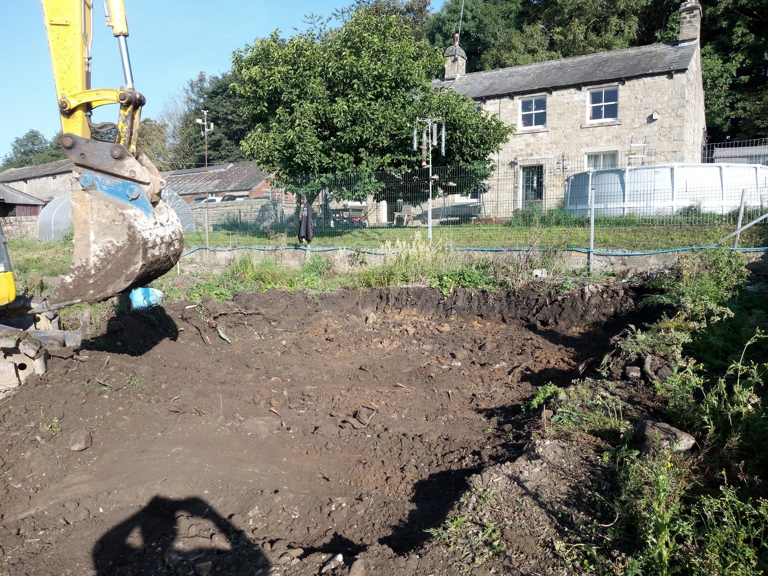 A construction site with an excavator bucket in the foreground, showing a cleared dirt area, house, trees, and a lush garden in the background.