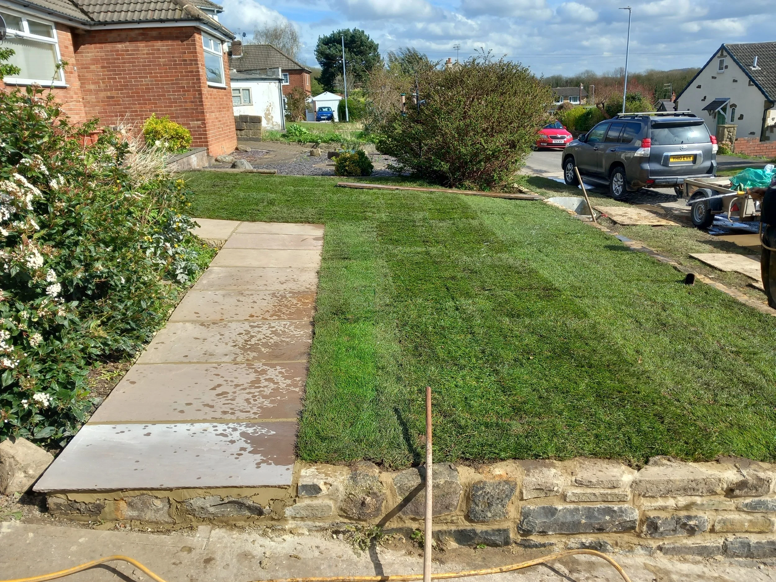 A residential front yard with freshly laid concrete slabs on the sidewalk and lawn, a hedge on the left, and a parked car in the driveway on a sunny day.