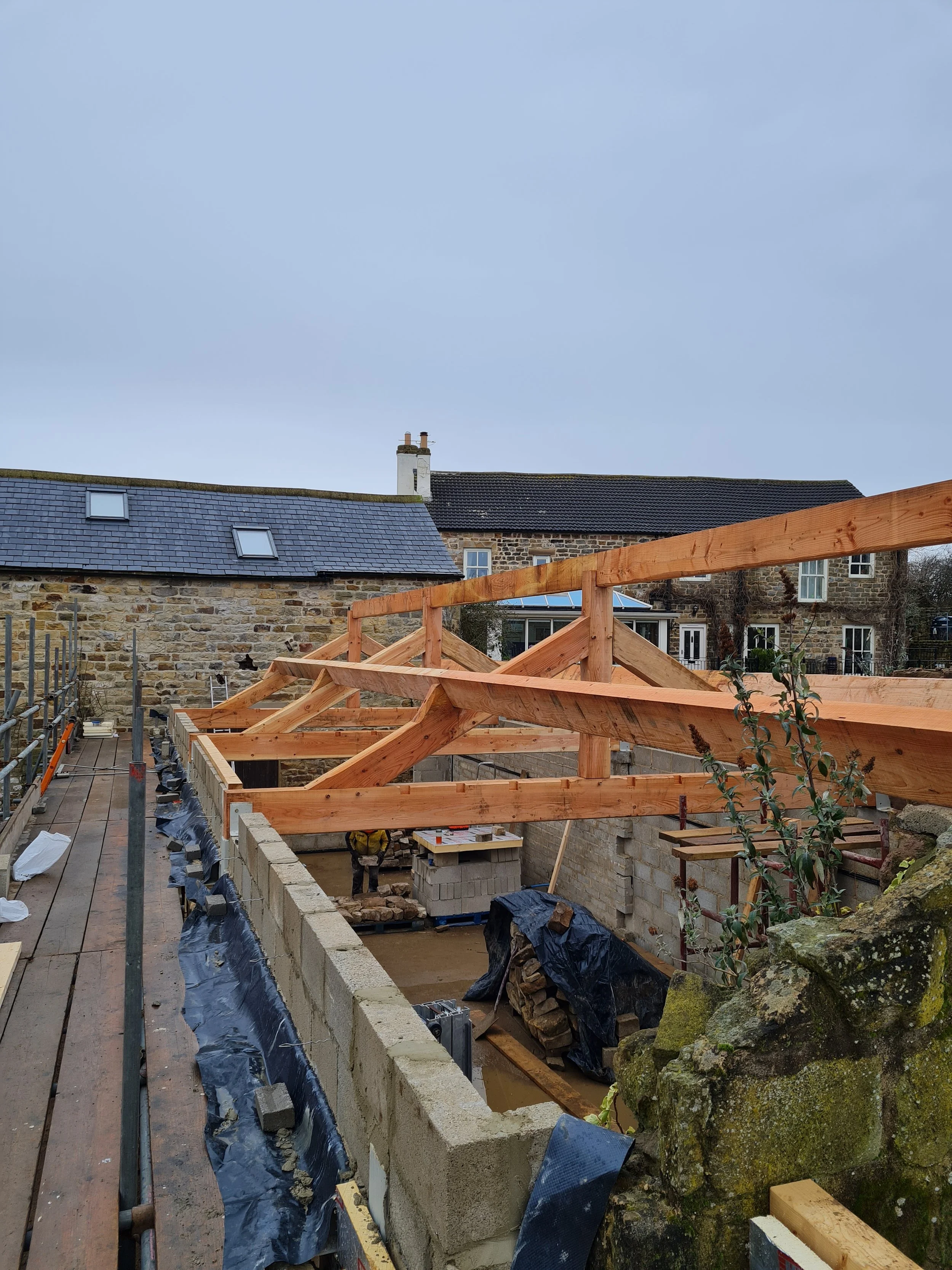 View of a construction site showing a partially built wooden roof structure over a stone wall, with some tools and construction materials, and neighboring stone buildings in the background.