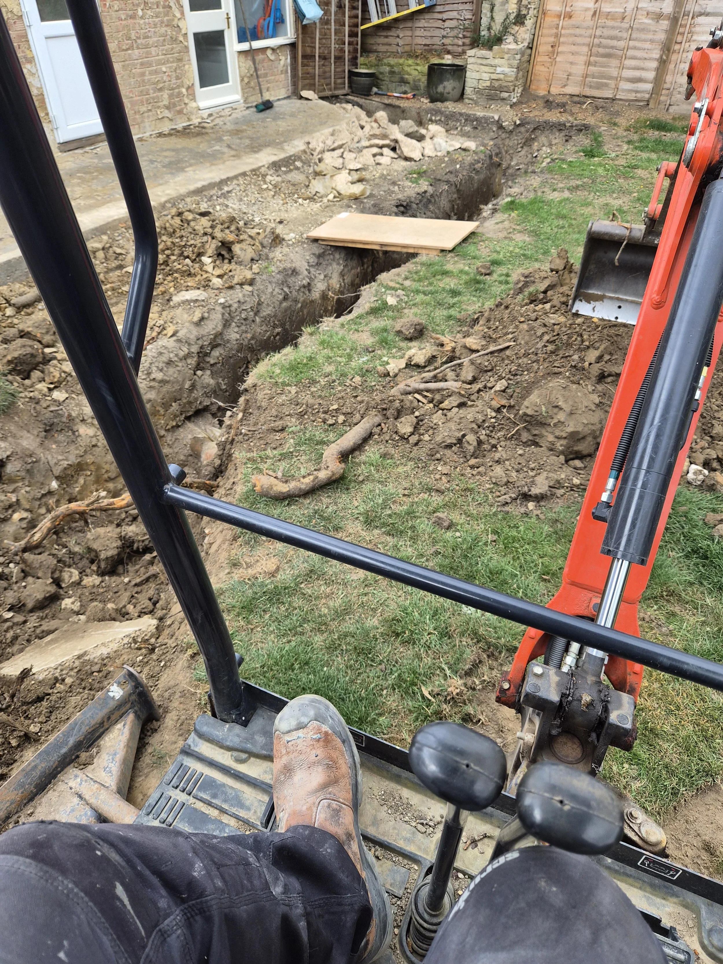 Construction site with excavator and trench dug in backyard, with dirt, rocks, and a wooden plank.