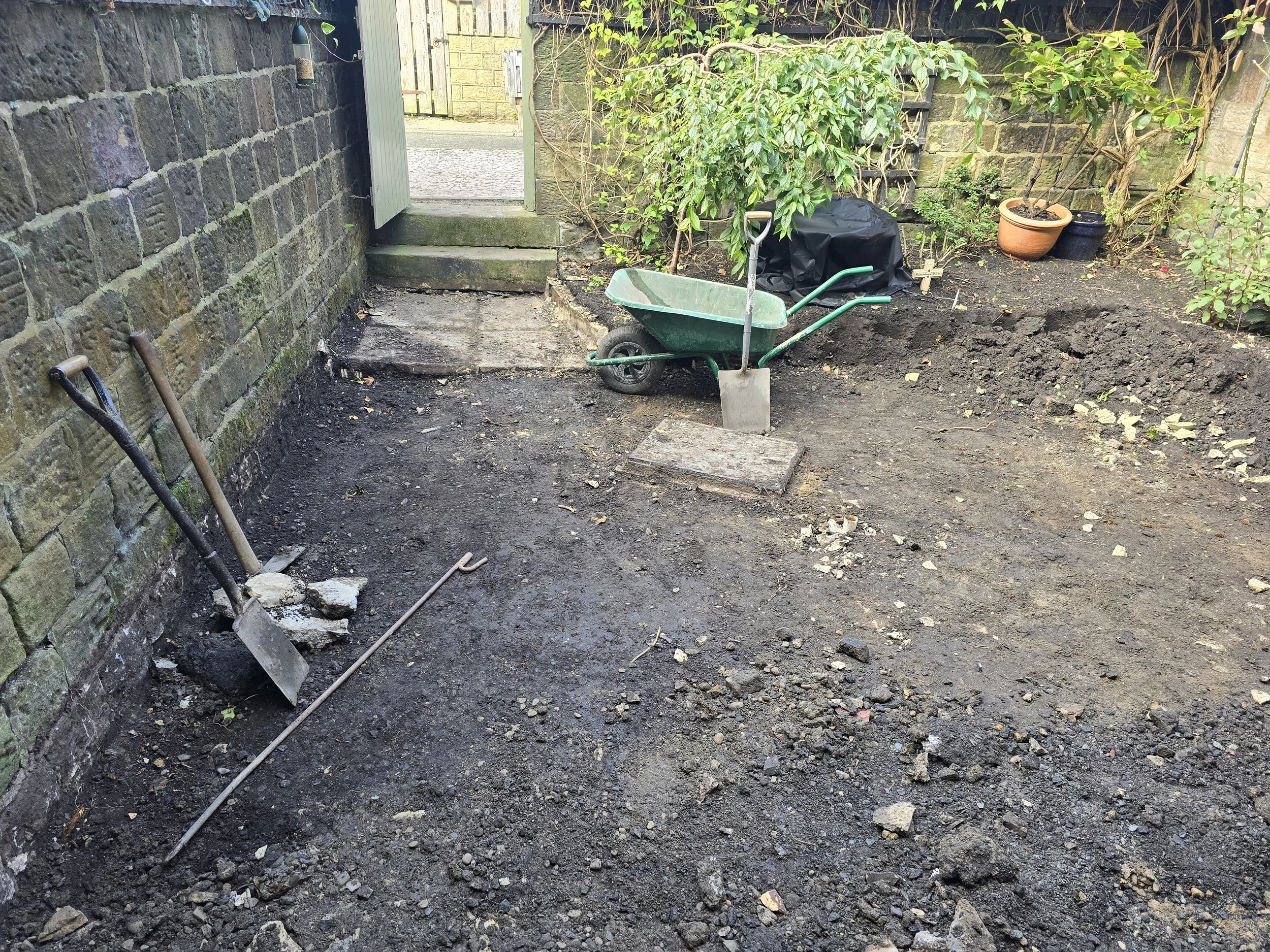A backyard with a partially dug soil area, gardening tools, a wheelbarrow, and potted plants near a brick wall and a gate.