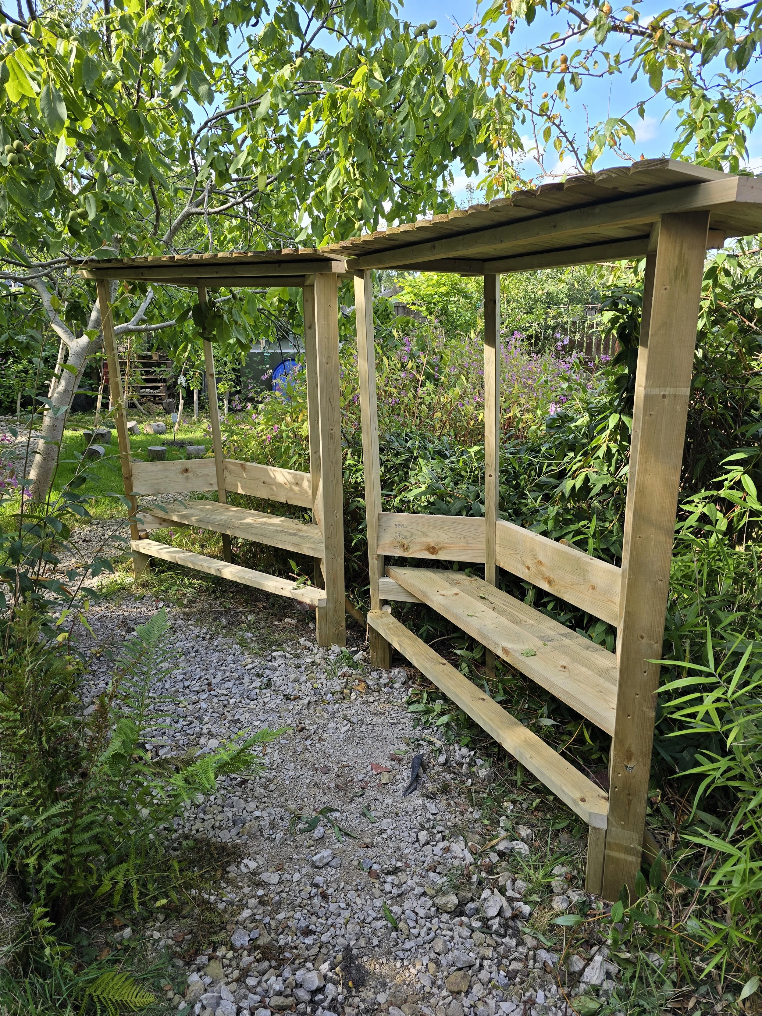 Unfinished wooden garden bench with two shelves on a gravel path, surrounded by green plants and trees under a blue sky.