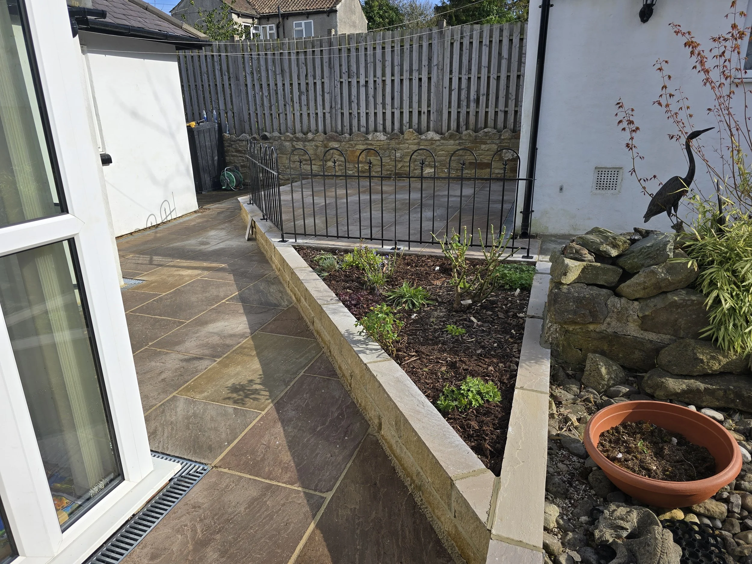 A backyard patio with large stone tiles, a raised flower bed with plants and shrubs, a decorative metal bird sculpture on rocks, a terracotta plant pot, and a black fence surrounding a small paved area, with a wooden fence and houses in the backgroun