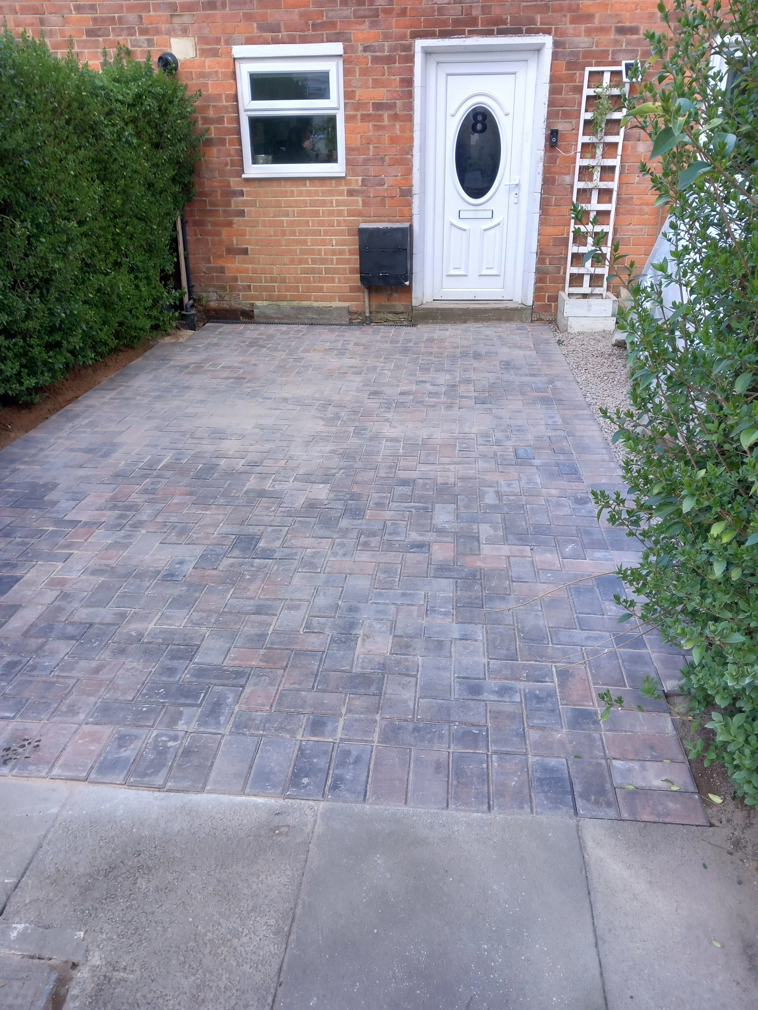A brick house entrance with a white door numbered 8, a window above, a black mailbox, some greenery on the left and right sides, and a paved driveway with grey and brown bricks.