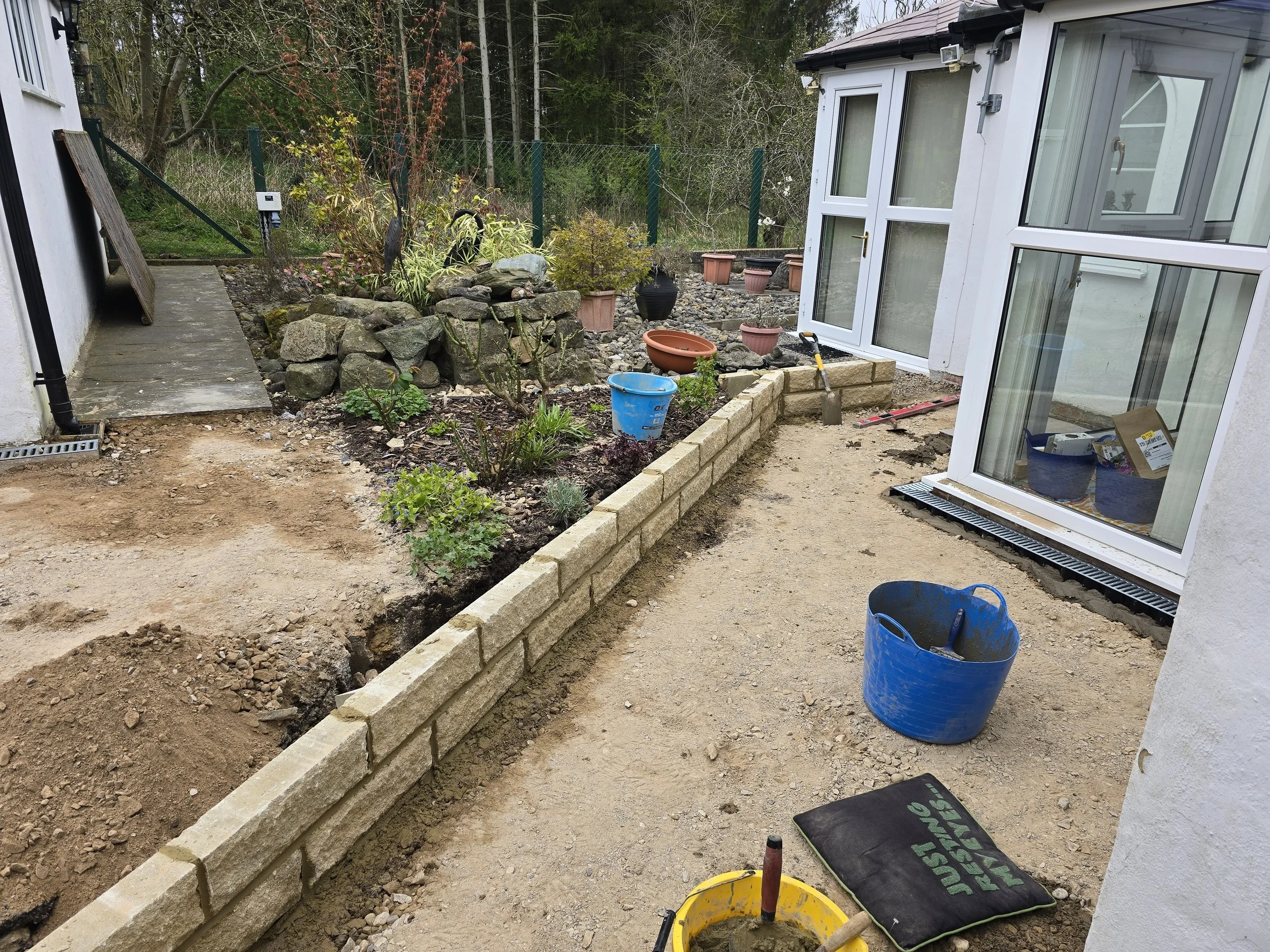 A backyard garden at the beginning stages of landscaping with a small stone wall being built along the house. There are garden plants, pots, and landscaping tools visible. A gravel path is outlined, and a pile of dirt and construction materials are i