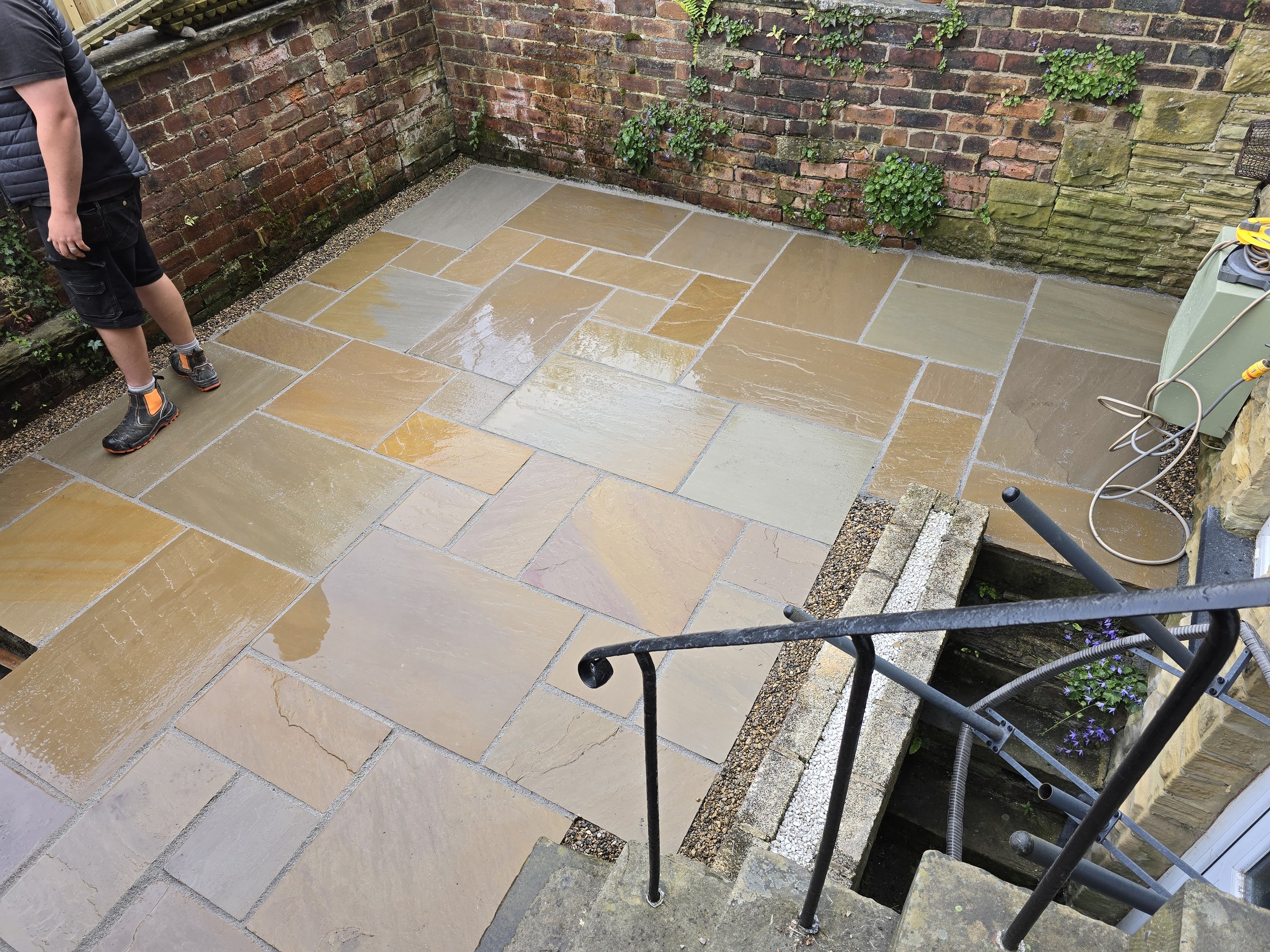 Wet stone patio with large beige, tan, and gray tiles, surrounded by brick walls and some green plants, viewed from above with part of a person's legs visible.