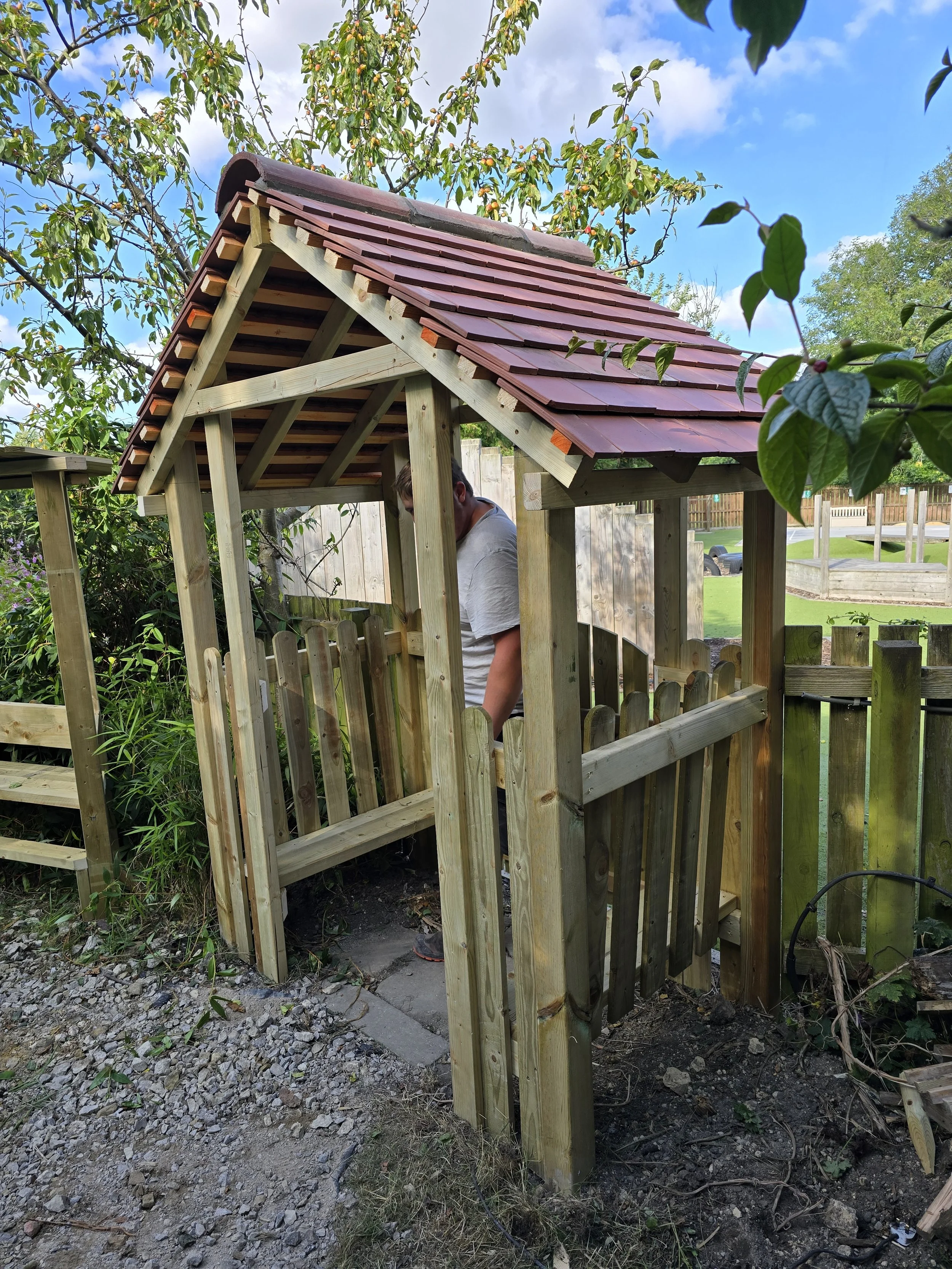 A person working on building a wooden playhouse or shed in a backyard, with trees and a fenced yard in the background.