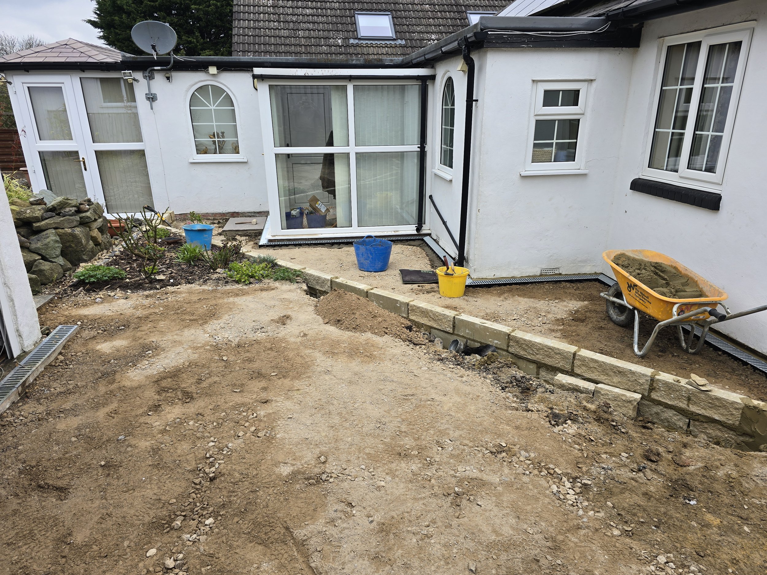 Backyard under renovation with dirt ground, construction tools, and partially built stone wall, attached to a white house with multiple windows and a glass conservatory.