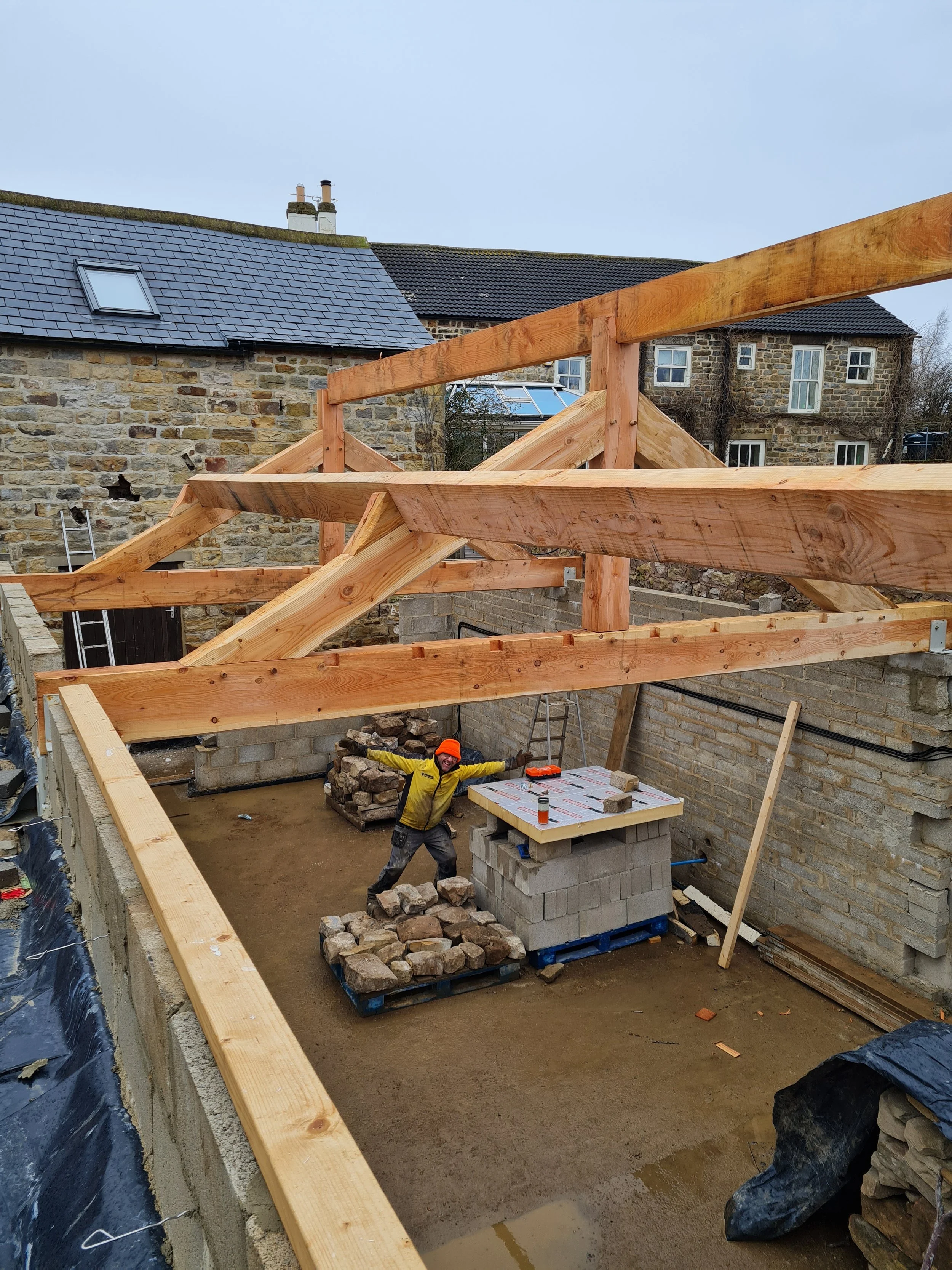 Construction worker wearing yellow jacket and orange helmet in a partially built structure with stone walls, wooden beams, and construction materials.