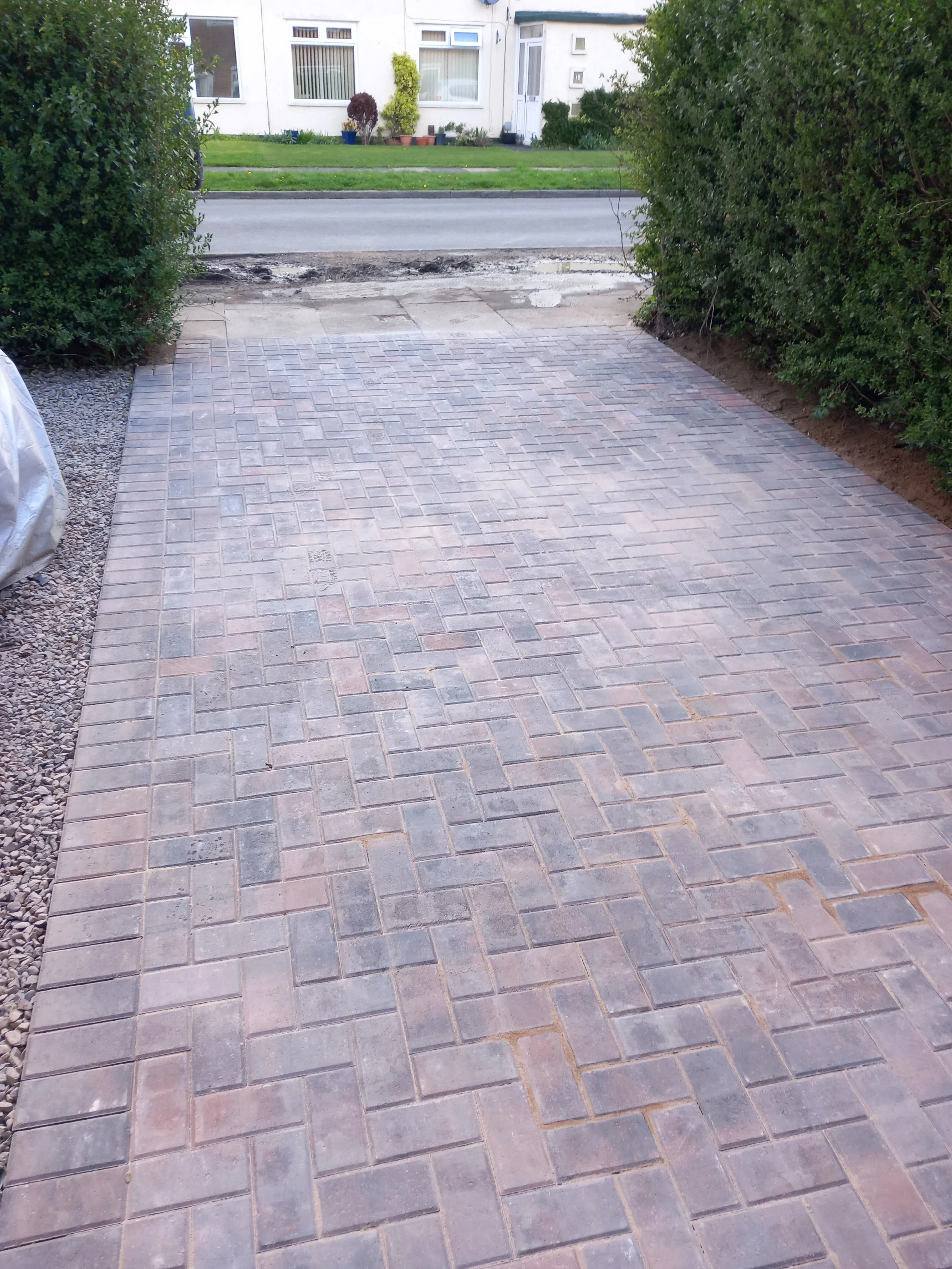 Newly paved brick driveway leading to a street with a house in the background, flanked by green hedges.