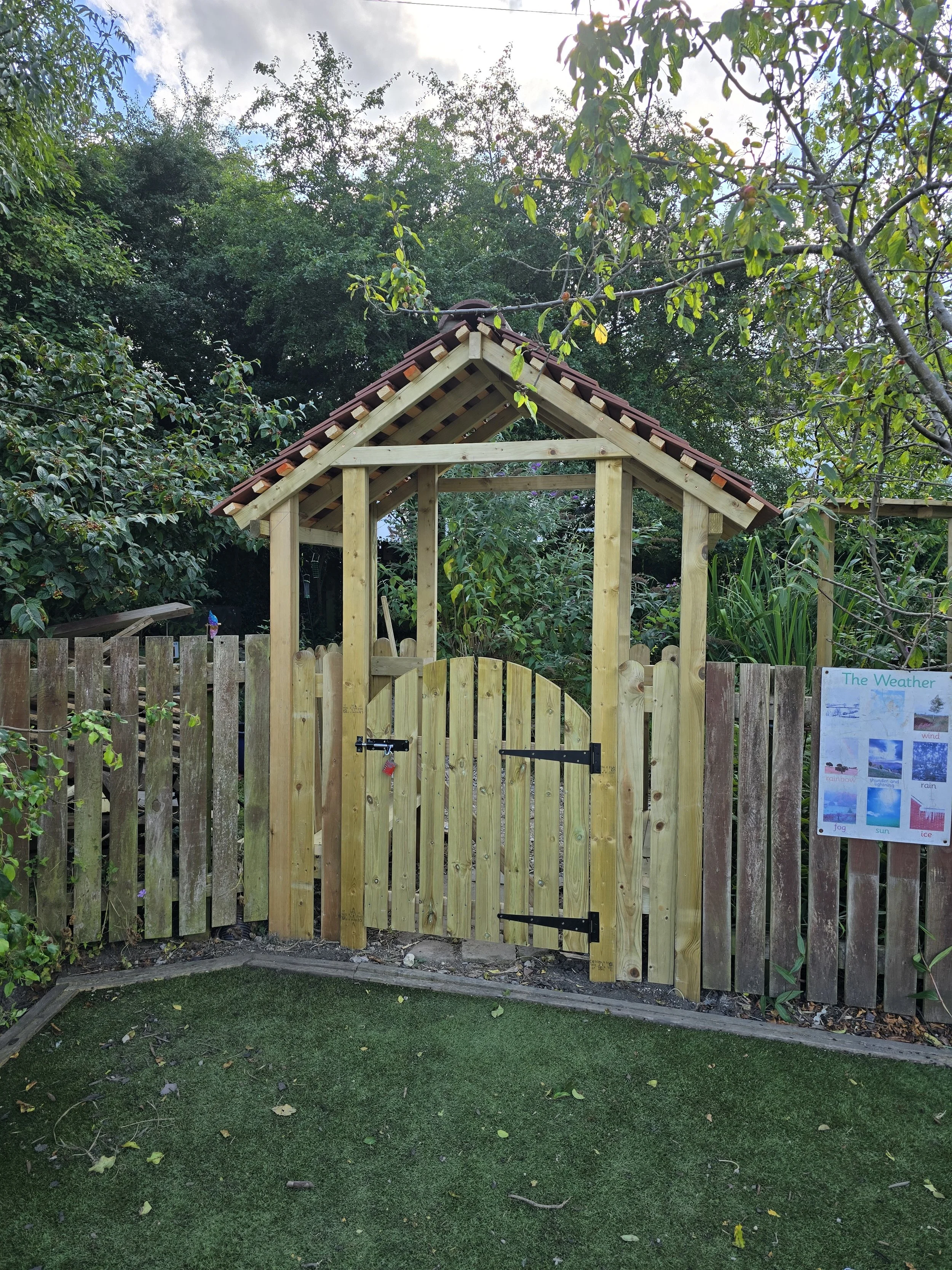 A newly built wooden garden gate with a small door, set in a wooden fence, under a red tile roof, surrounded by trees and plants, with an educational weather poster on the right side.