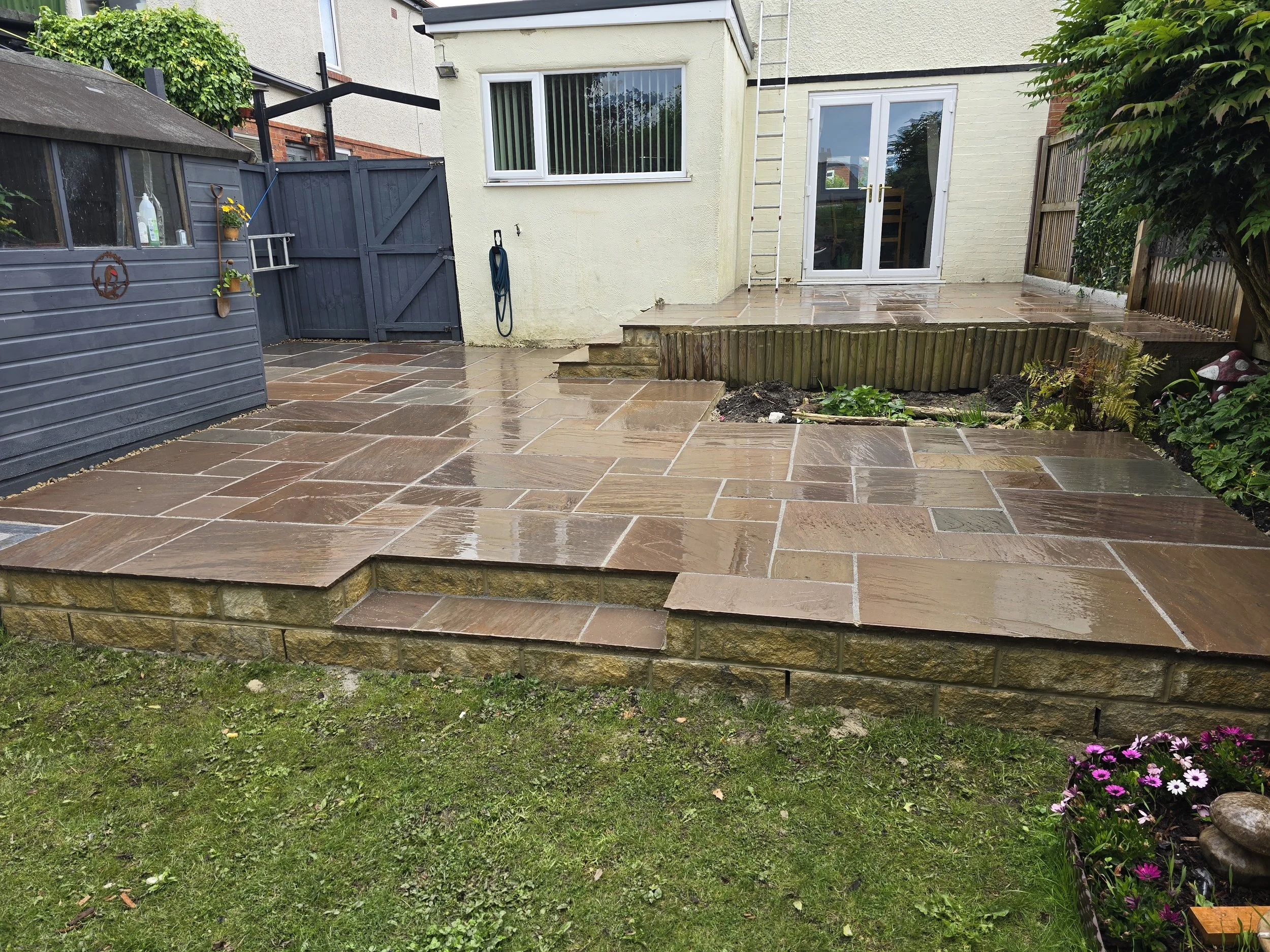 Backyard patio with wet brown stone tiles, a small garden bed, a blue shed on the left, and a white house with a sliding glass door in the background.