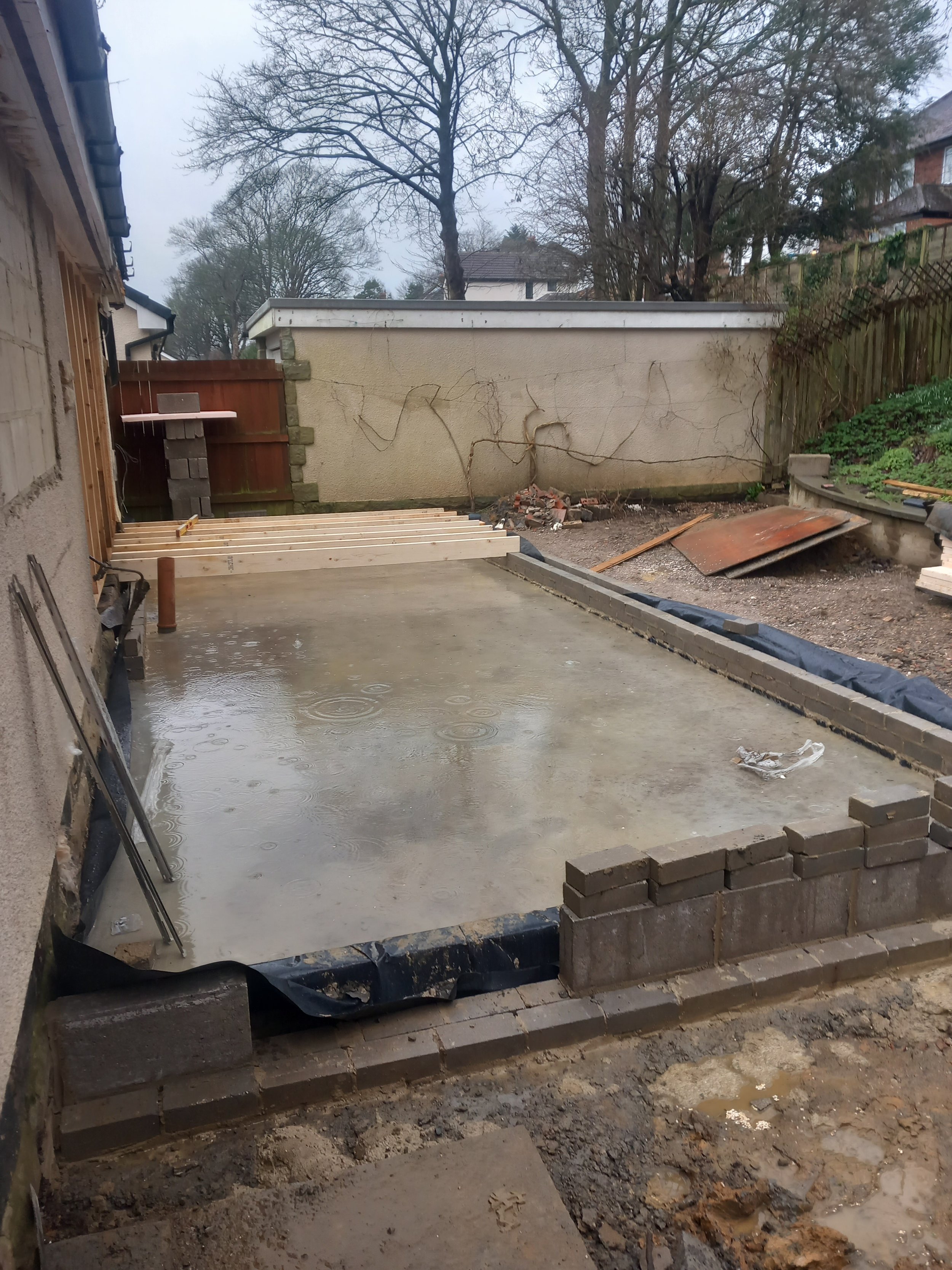 Construction site with a concrete foundation and partially built brick wall, adjacent to a house with construction tools and materials scattered around, on a rainy day.
