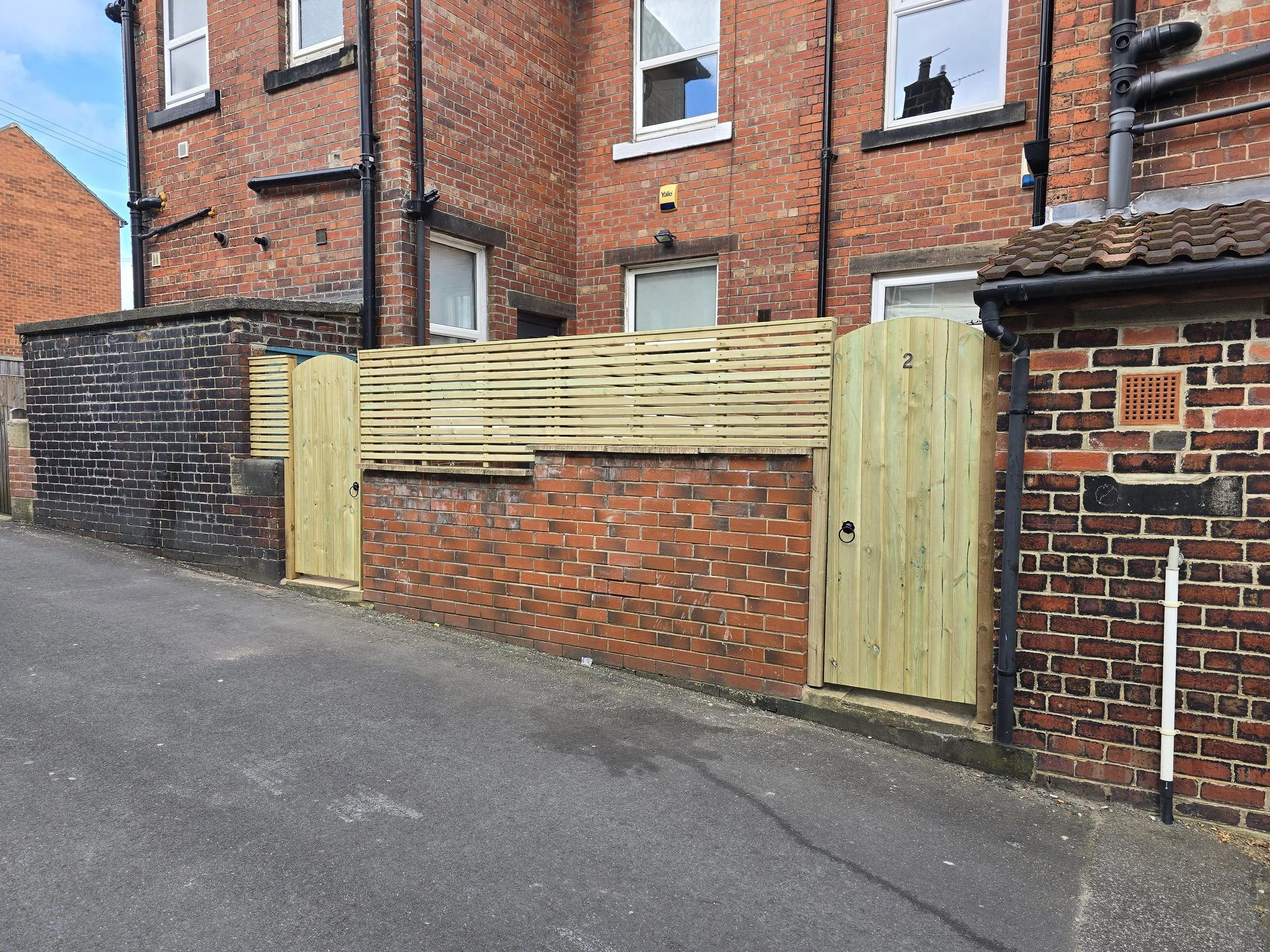 Newly constructed wooden gate and fence in front of a brick building with multiple windows and black drain pipes.