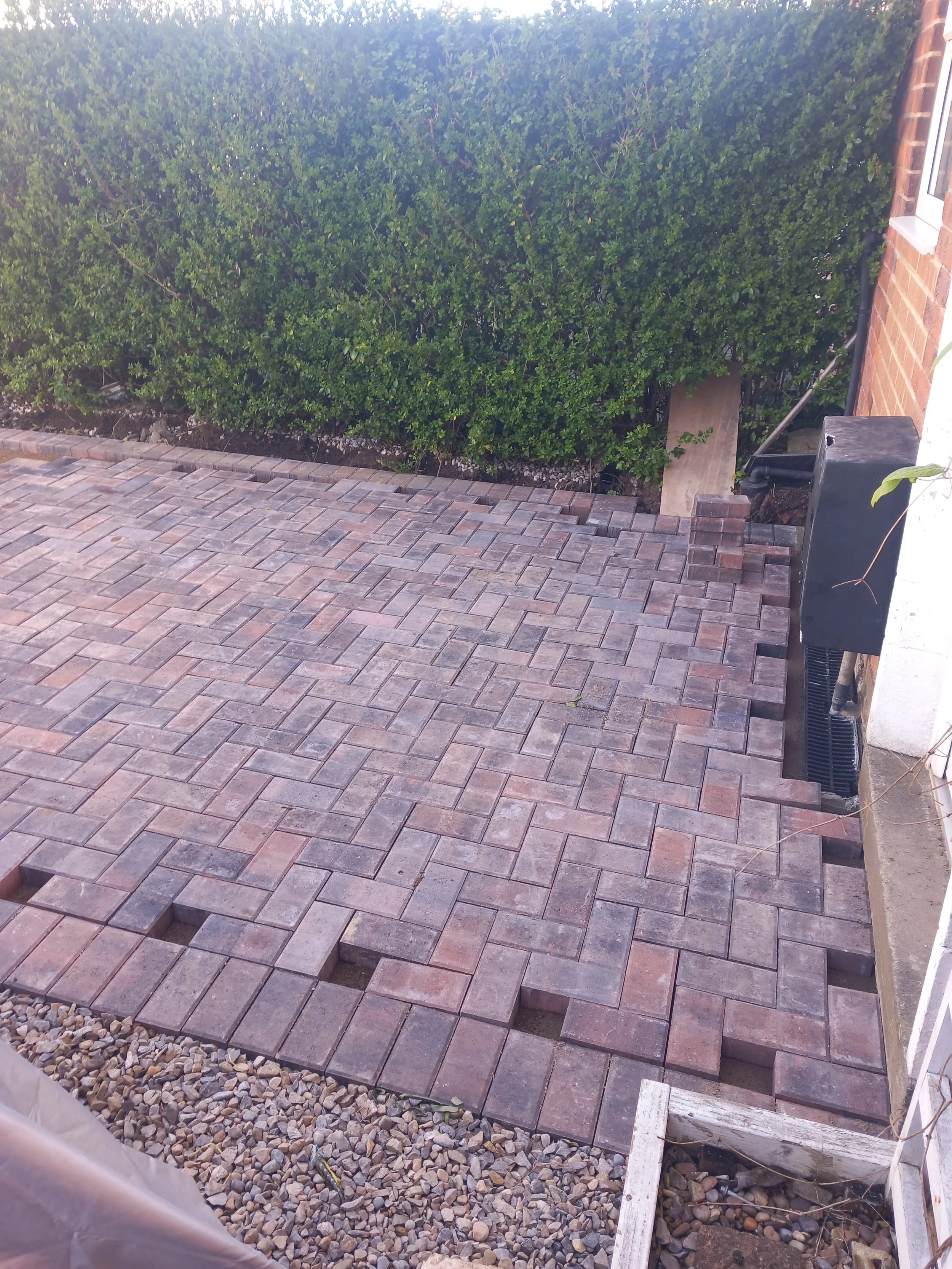 Newly paved brick patio area next to a brick house with a black utility box, a wooden plank, and a green shrub in the background.