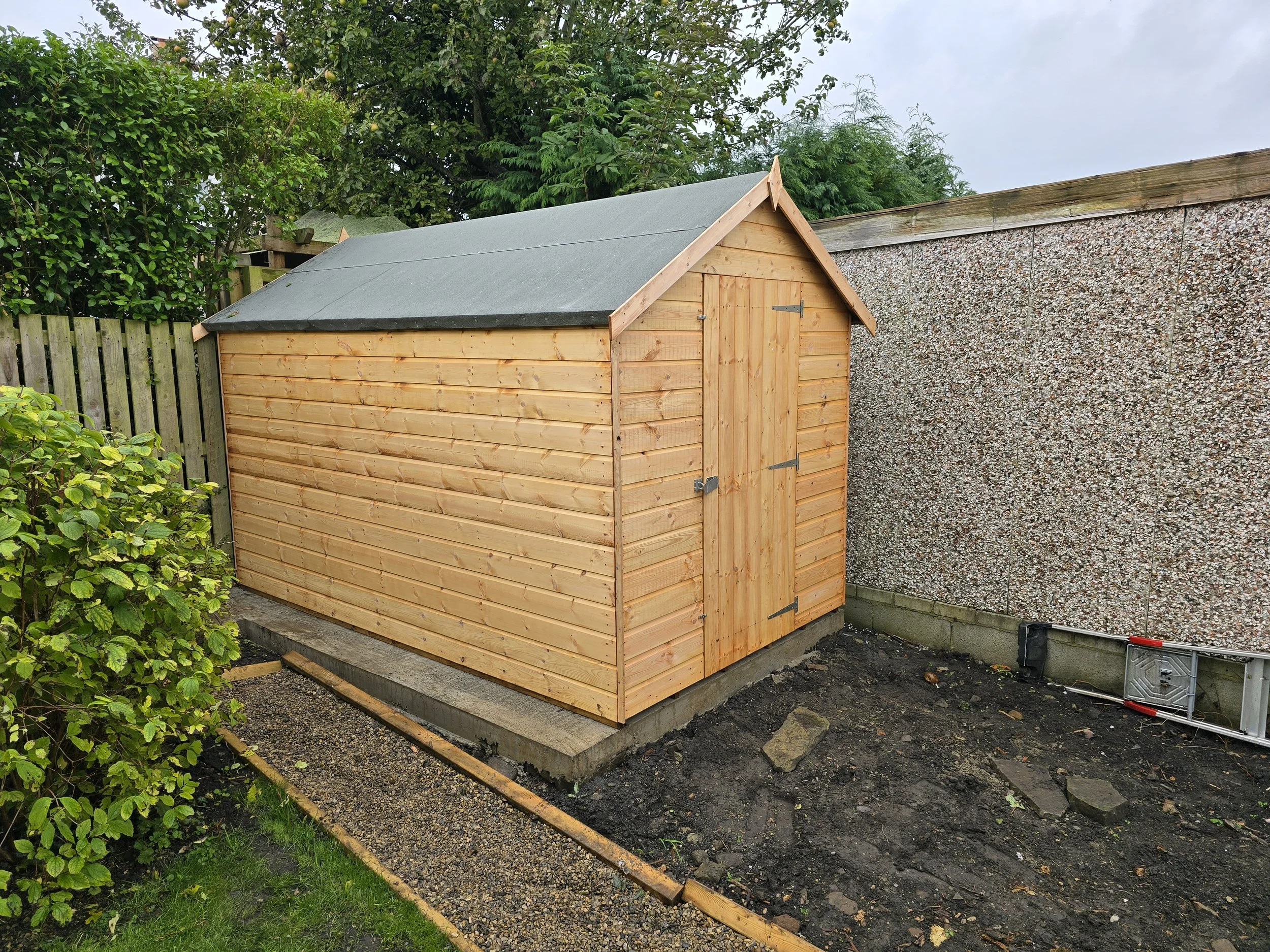 A newly built wooden shed with a pitched roof, situated in a backyard against a gravel wall. The shed has a wooden door secured with metal hinges. Surrounding the shed are a wooden fence, green bushes, and a patch of bare soil.