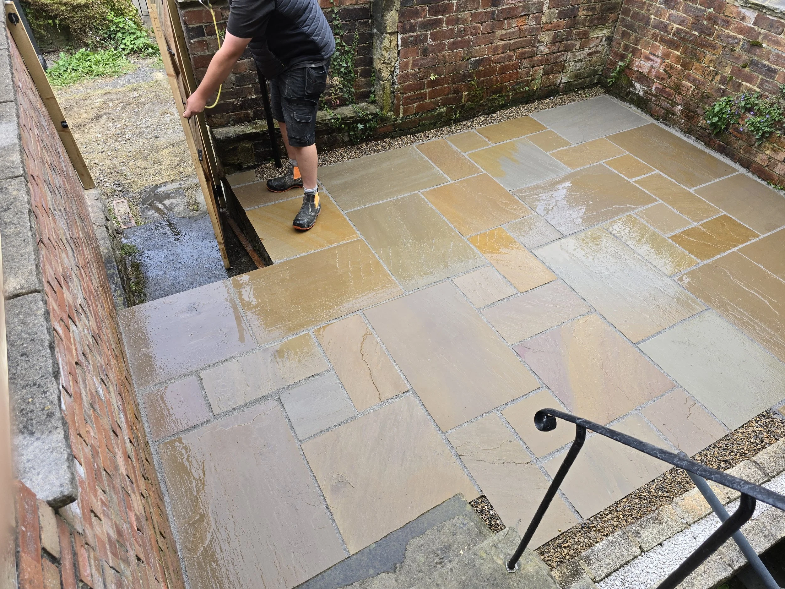 A person working on installing a stone patio surrounded by brick walls, with wet stones and some gardening tools nearby.