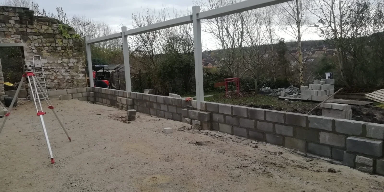 Construction site with a partially built brick wall, building materials, and a tripod measuring tool on sandy ground in an outdoor area with trees and a cloudy sky.
